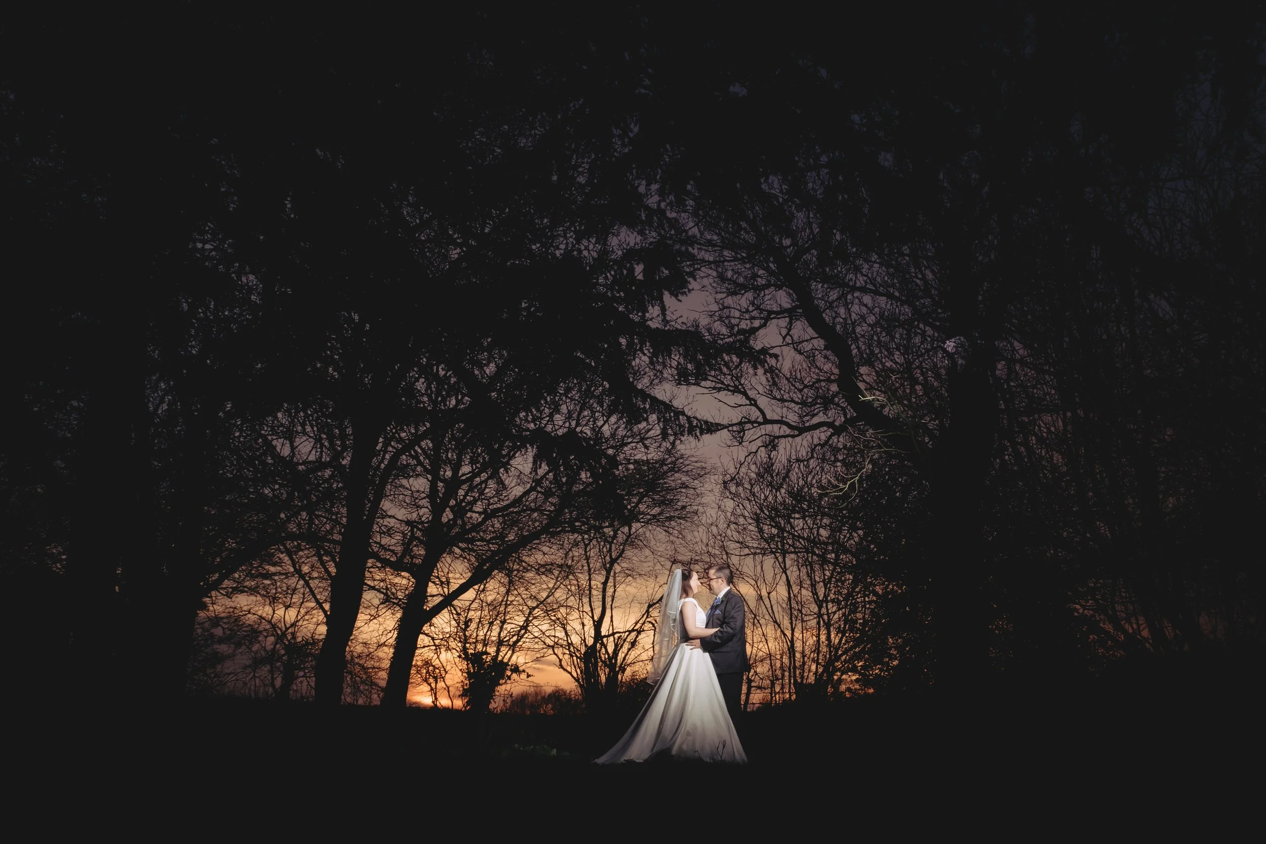 A bride and groom standing close together holding hands in a wooded area at sunset, with silhouettes of trees and a colorful sky in the background.