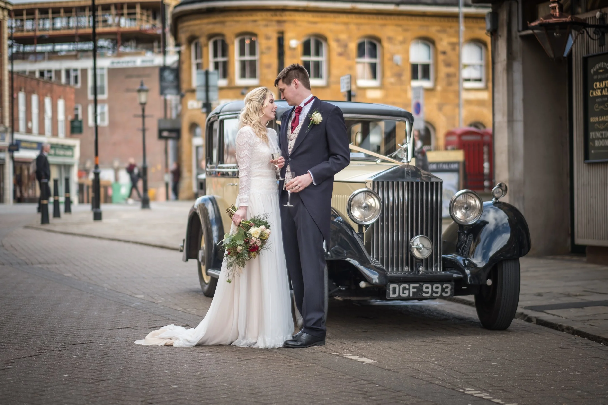 A bride and groom standing close together on a city street, gazing at each other, with the bride holding a bouquet and the groom holding a champagne glass, in front of a vintage car with a city backdrop.