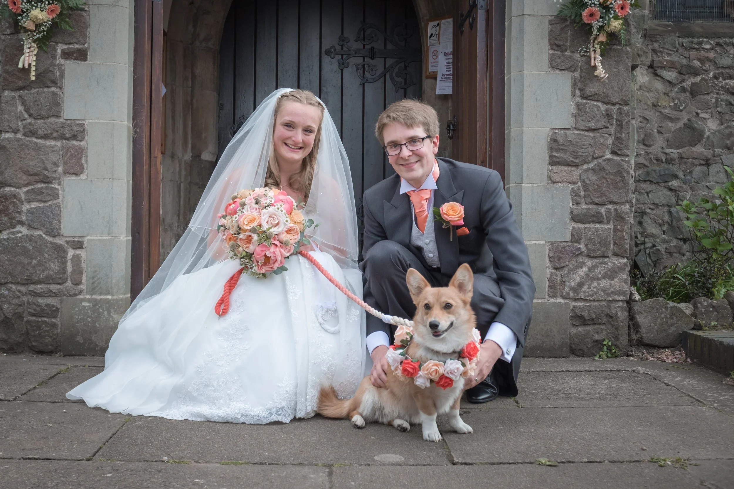 A bride and groom crouching together in front of a stone building entrance, smiling at the camera with a dog in a flower collar in front of them.