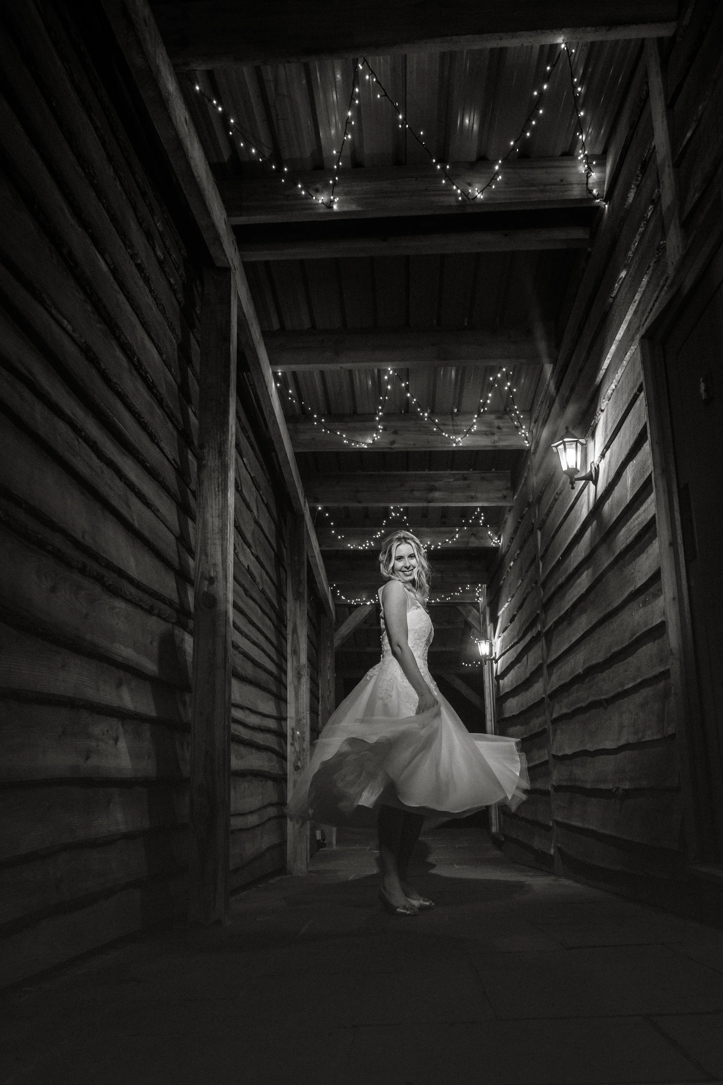 Black and white photo of a woman in a wedding dress standing in a dimly lit wooden corridor with string lights overhead and a lantern on the wall.