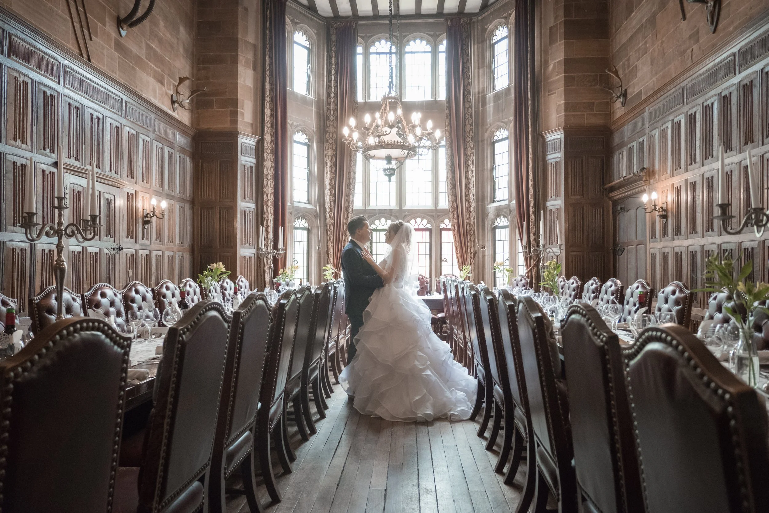 A bride and groom are dancing in a grand, historic banquet hall with high stained glass windows, dark wooden walls, and ornate chandeliers.