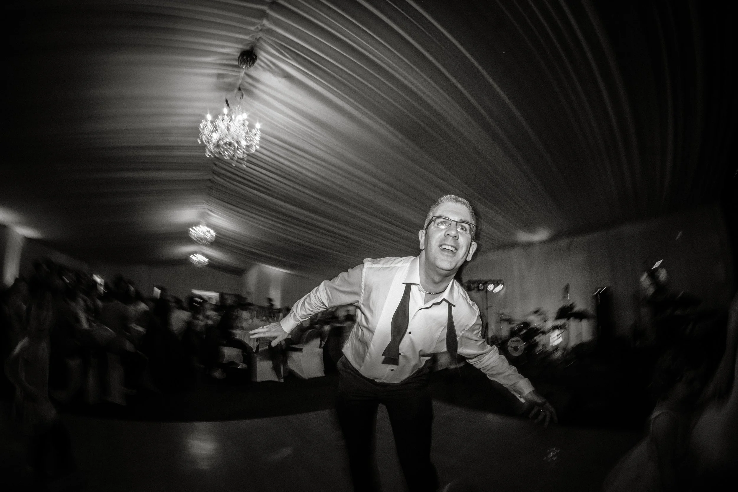 A man wearing glasses and a dress shirt with a loosened tie, smiling and leaning forward in a dance or performance setting, with a chandelier and curtains visible in the background, black and white photo.