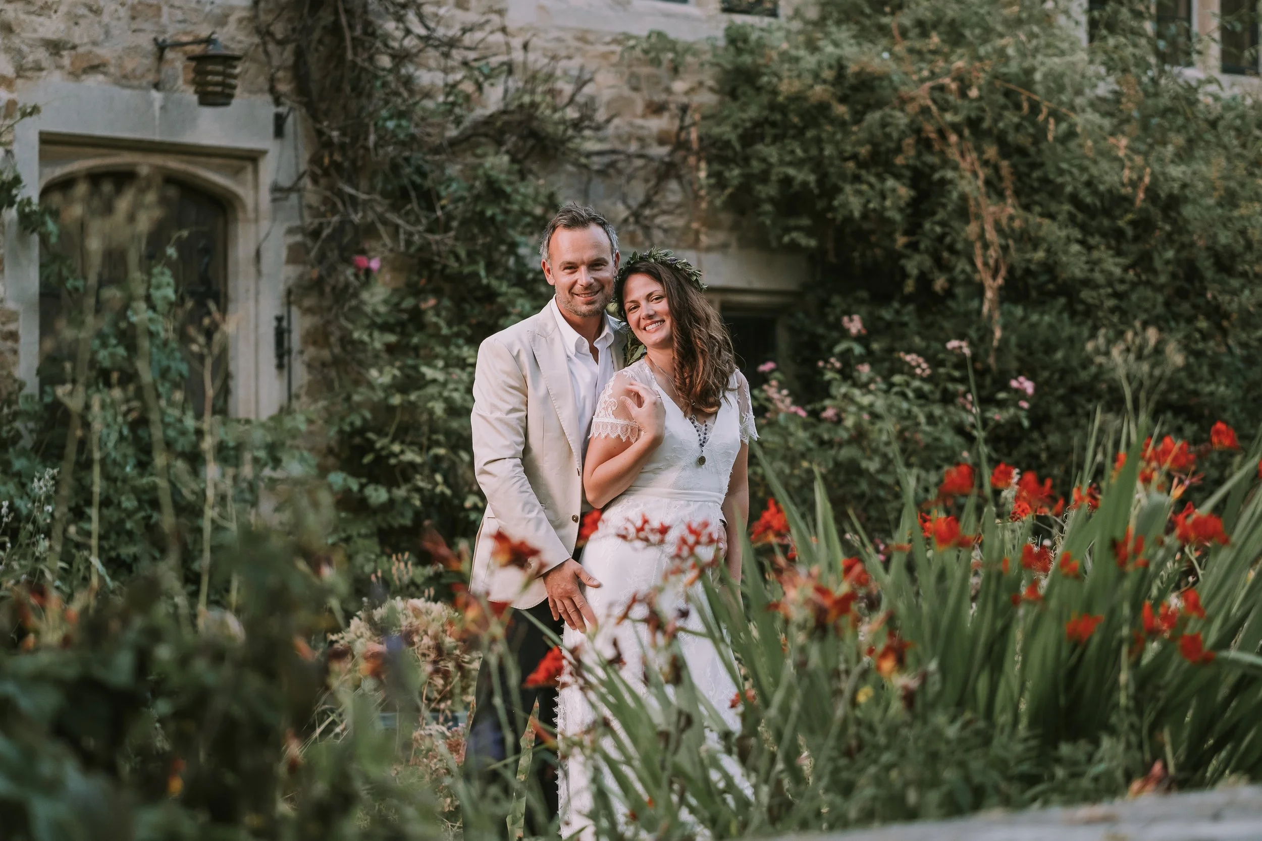 A couple in wedding attire stands arm in arm in a lush garden with blooming flowers and greenery, with a stone building in the background.