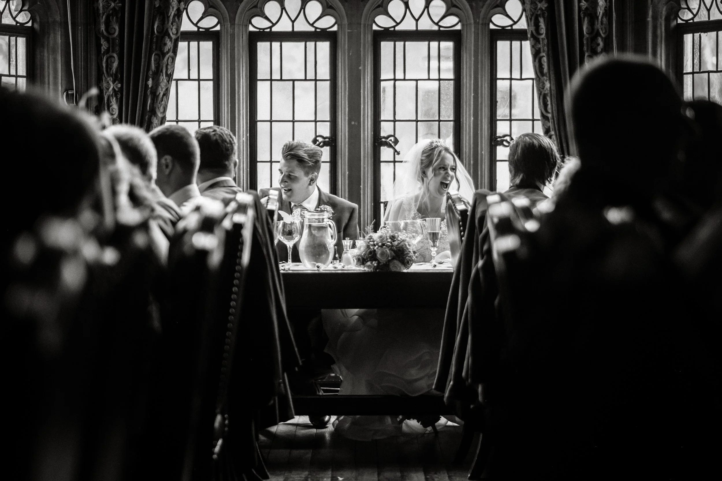 Black and white photo of a wedding reception in a historic building with tall, ornate windows and curtains. The bride and groom are seated at the head table, smiling and sharing a joyful moment. Photographers and guests are blurred in the foreground.