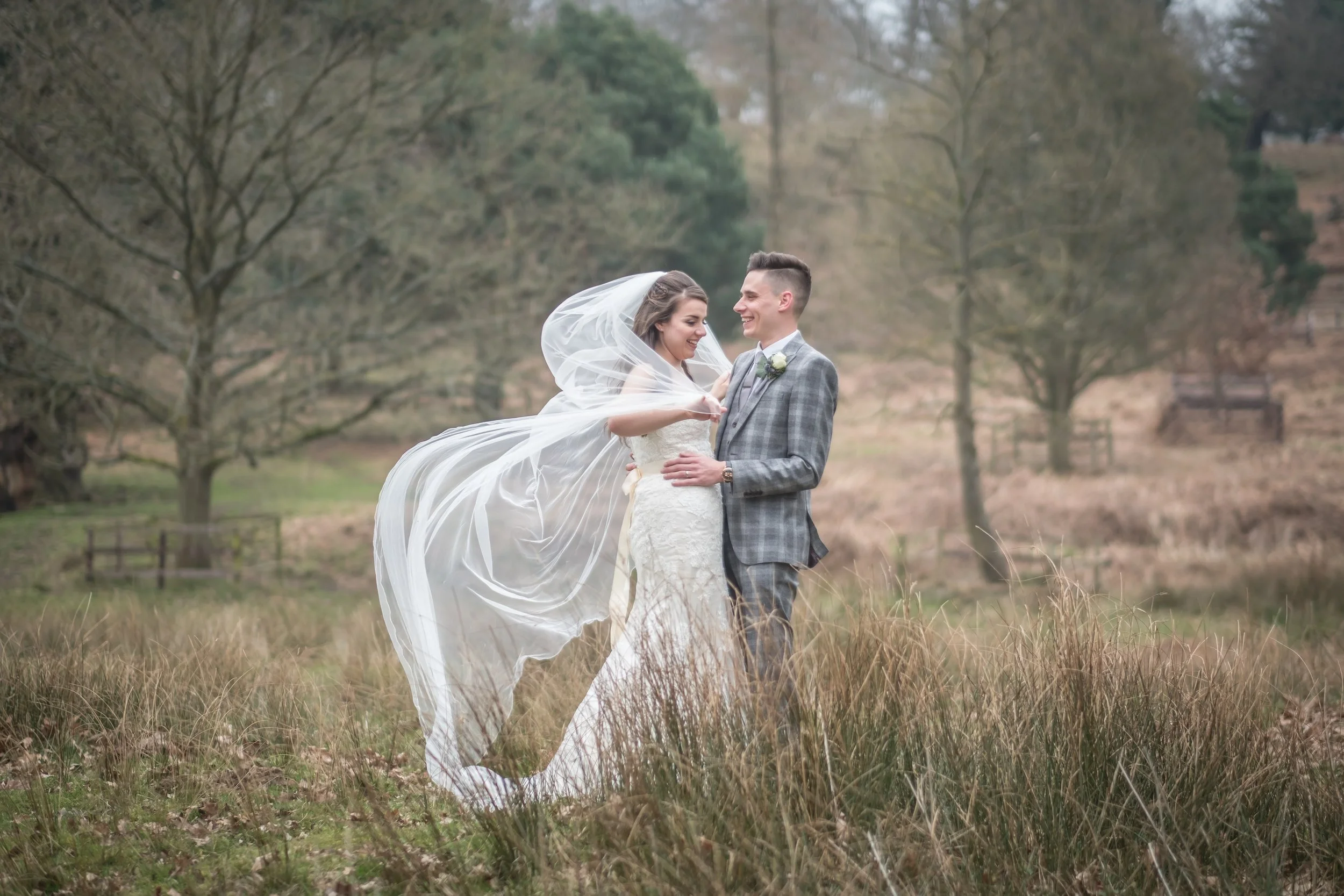 A bride and groom standing in a field with tall grass, surrounded by trees, during an outdoor wedding. The bride is holding the groom's tie and wearing a white dress with a veil, while the groom is wearing a gray checked suit. They are smiling at eac