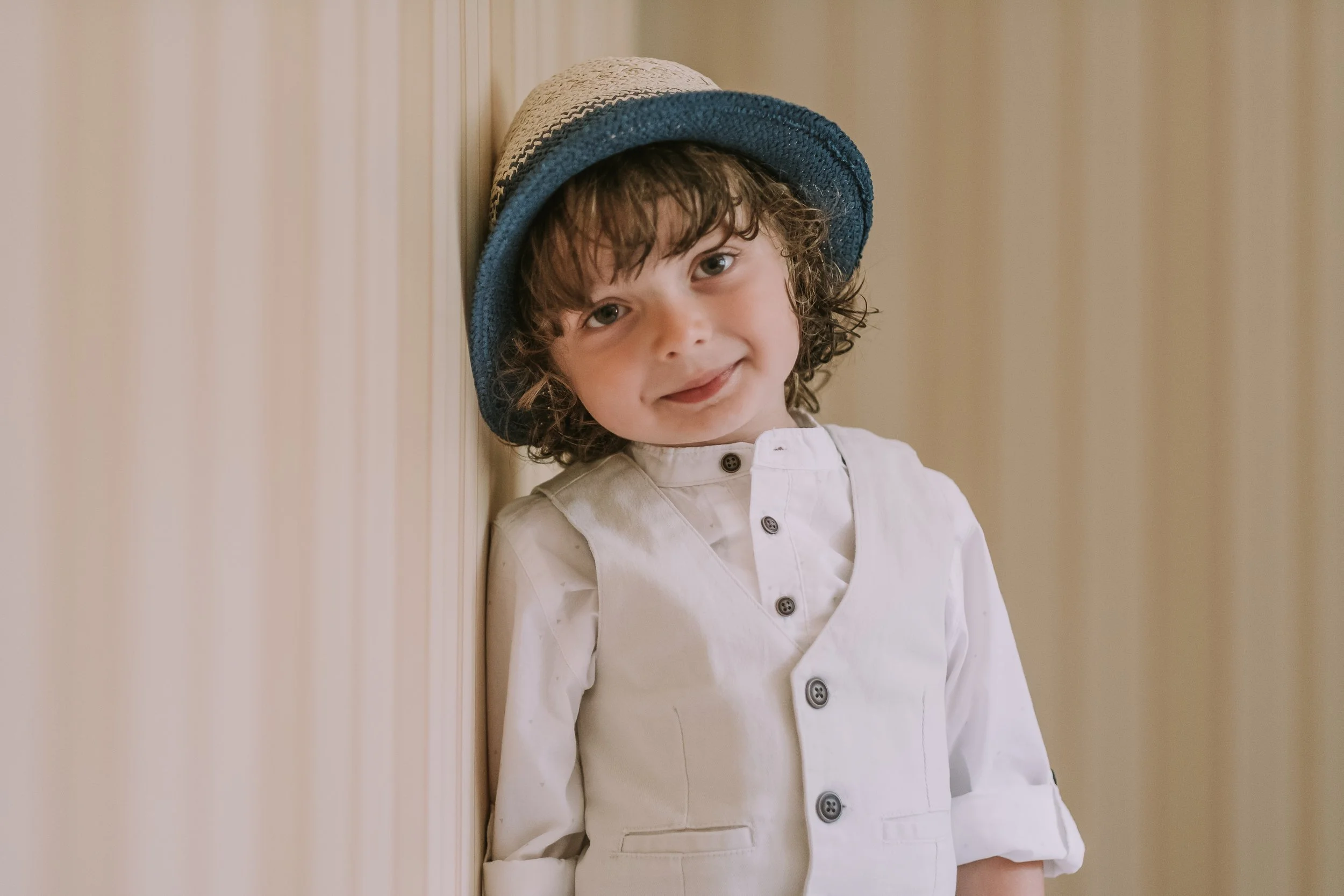 A young boy with curly hair wearing a beige and blue hat and a white jacket smiling while standing against a light-colored wooden wall.