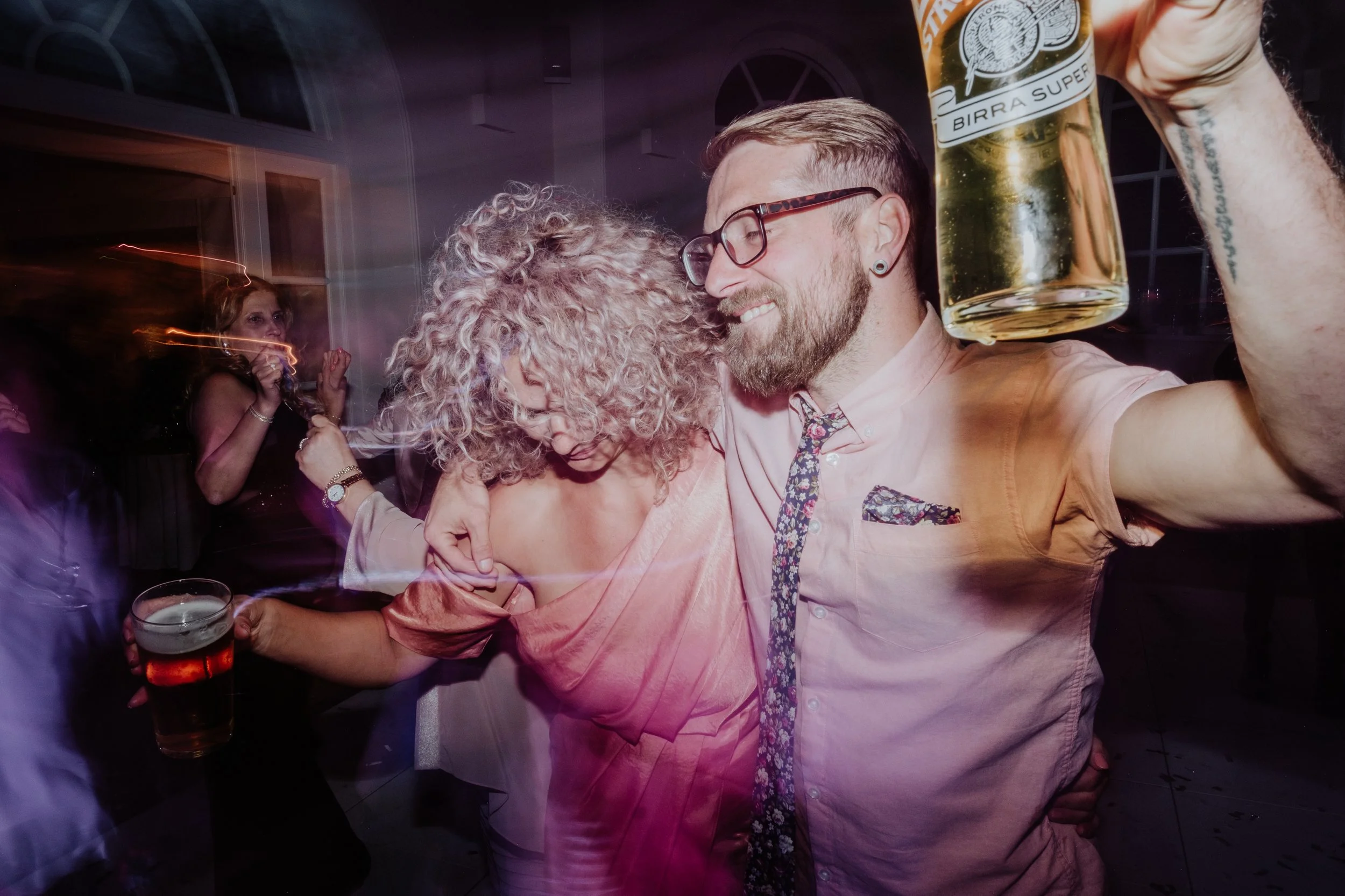 Young man with glasses holding a beer mug, smiling, dancing with a woman with curly hair at a party.