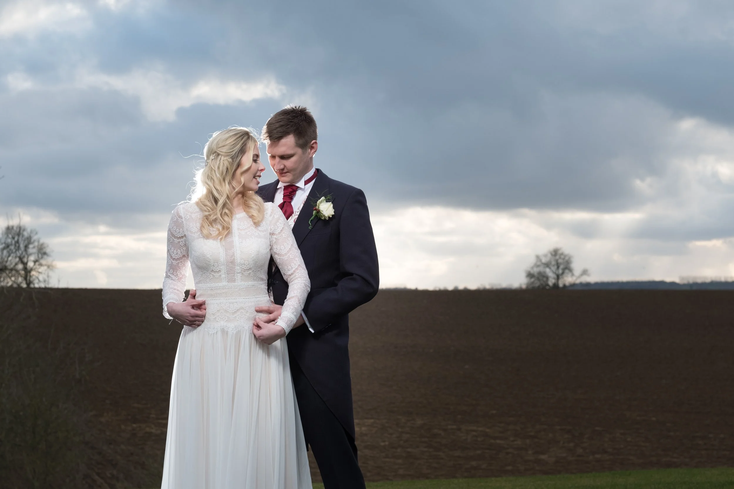 A bride and groom standing close together outdoors, with dark clouds in the sky, in a rural landscape.