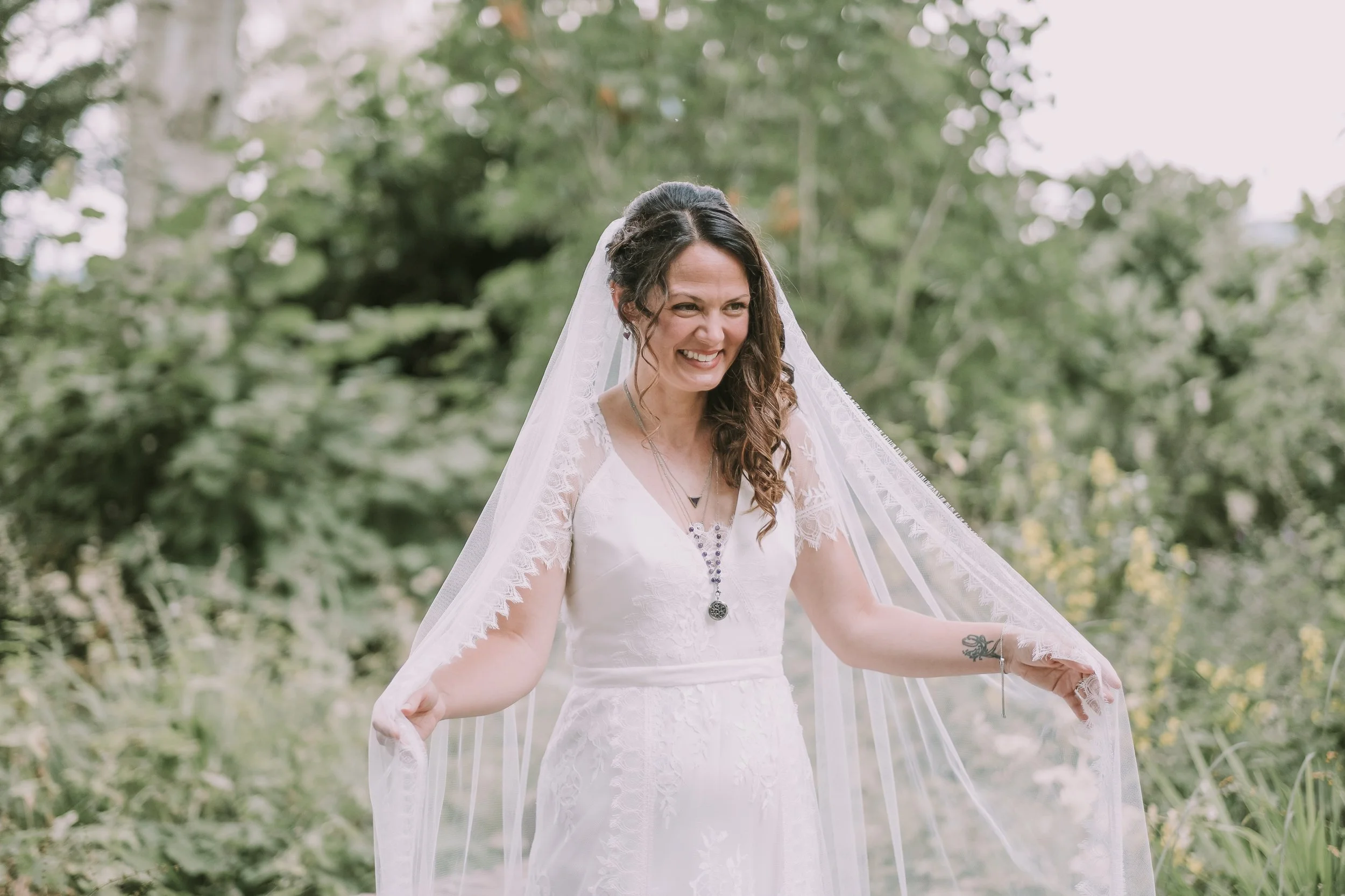 A woman in a white wedding dress and veil smiling outdoors amidst green foliage.