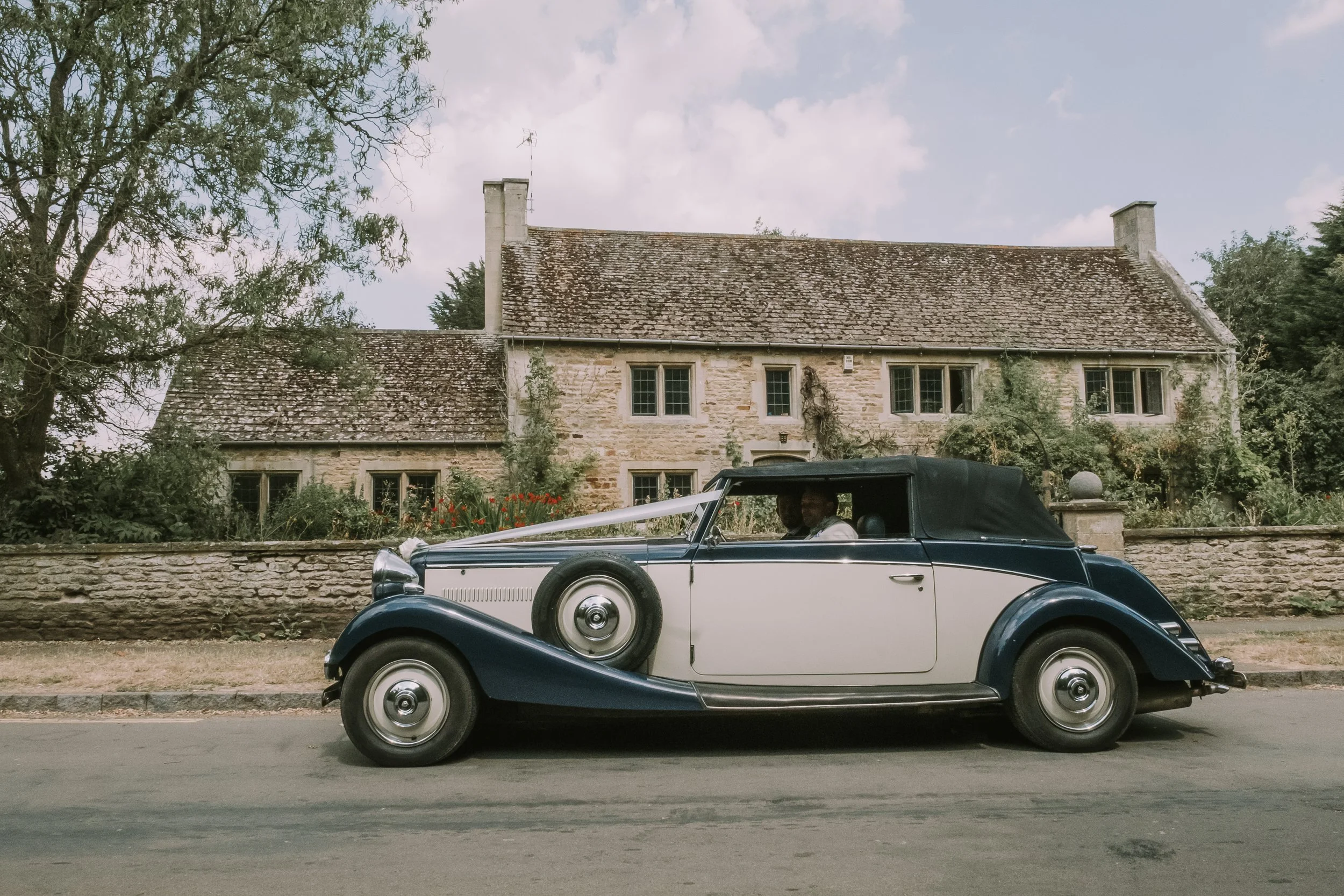 A vintage black and white convertible car with beige accents parked in front of an old stone house with lush greenery and flowers, under a cloudy sky.