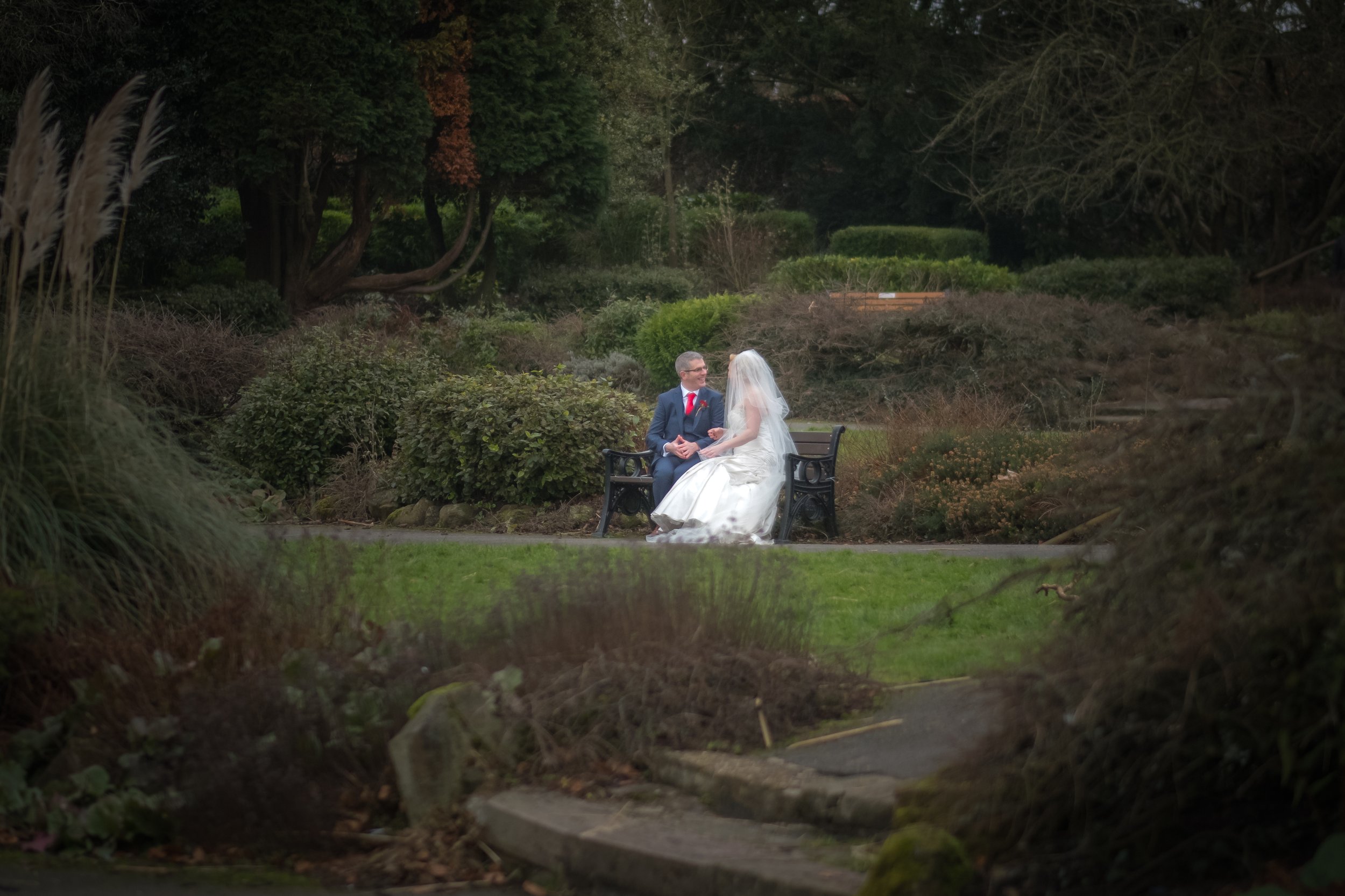 A bride and groom sitting on a park bench surrounded by greenery and trees, with a natural landscape background.