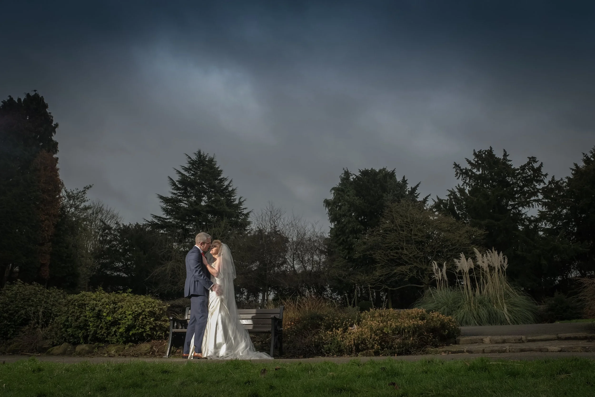 A bride and groom standing close together outdoors, holding hands, with trees and bushes in the background on a cloudy day.