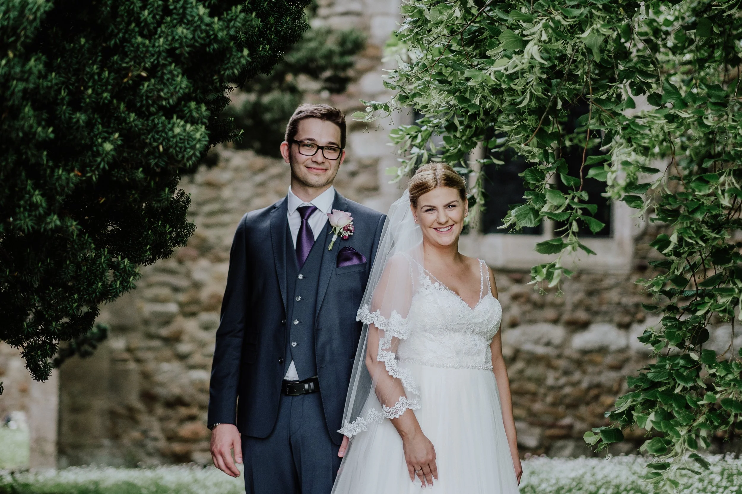 Bride and groom standing outdoors framed by green foliage during wedding.