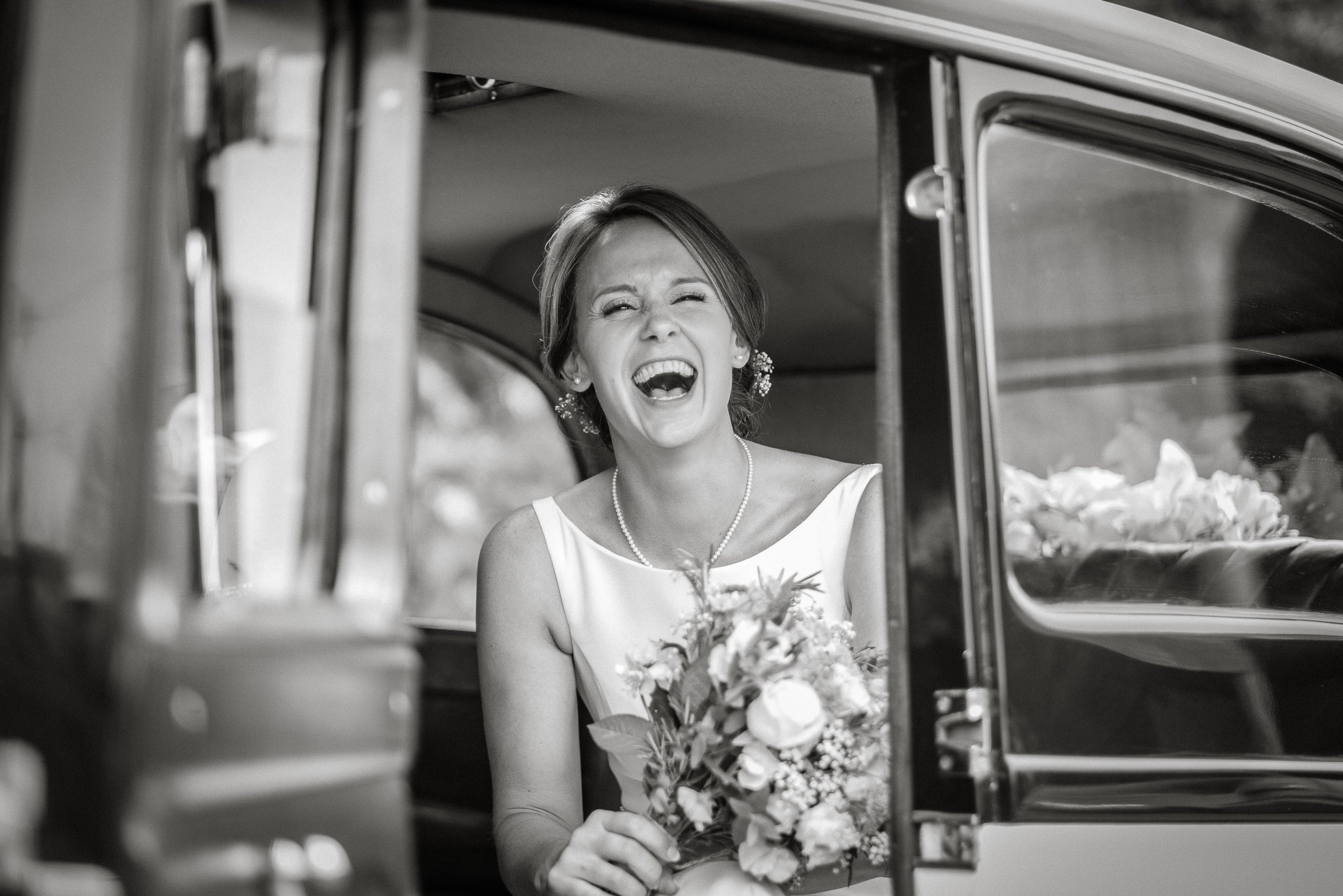 A woman dressed in a wedding gown laughing joyfully while looking out of a vehicle window. She is holding a bouquet of flowers and wearing earrings and a pearl necklace. The photo appears to be candid and is in black and white.