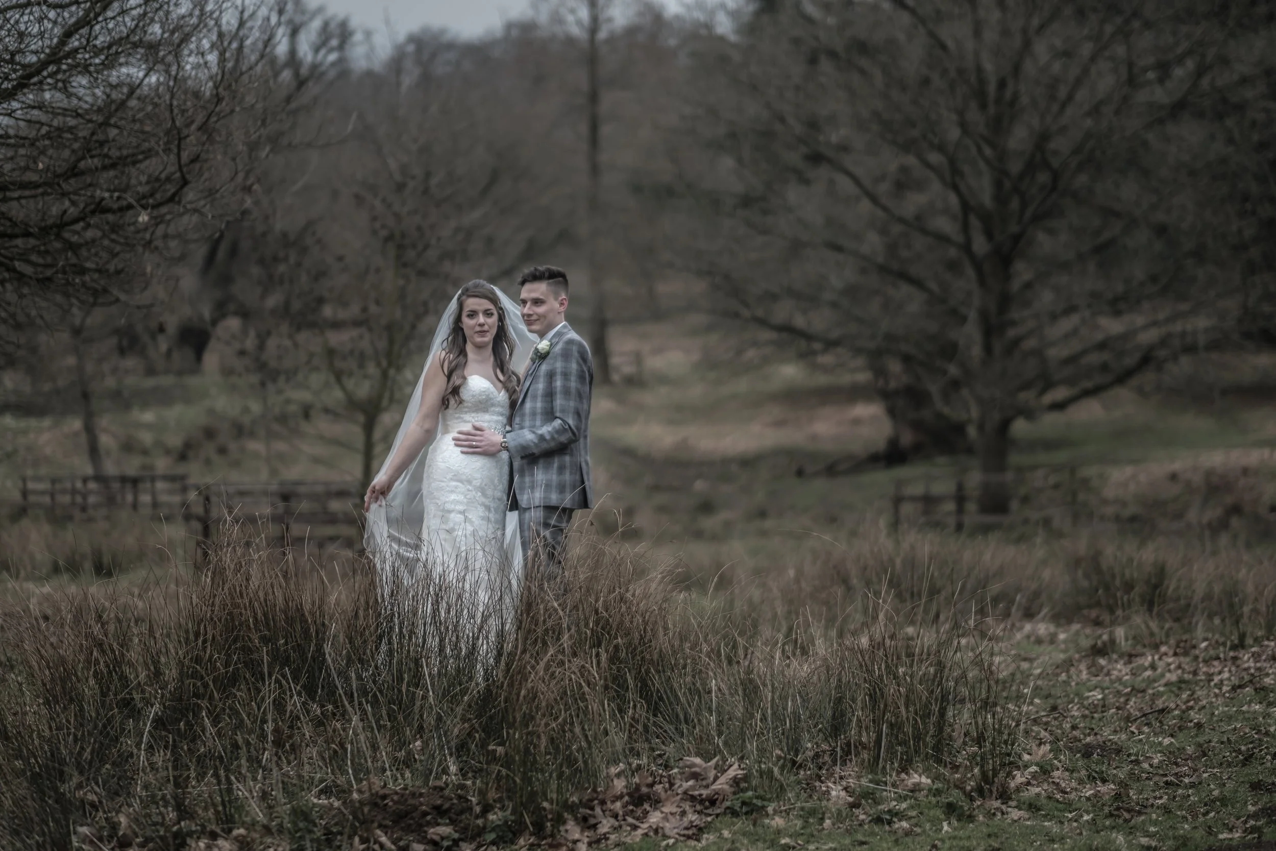 A bride and groom standing close together outdoors in a natural setting with leafless trees and grass, dressed in wedding attire, with the bride in a white lace gown and veil, and the groom in a gray checkered suit.