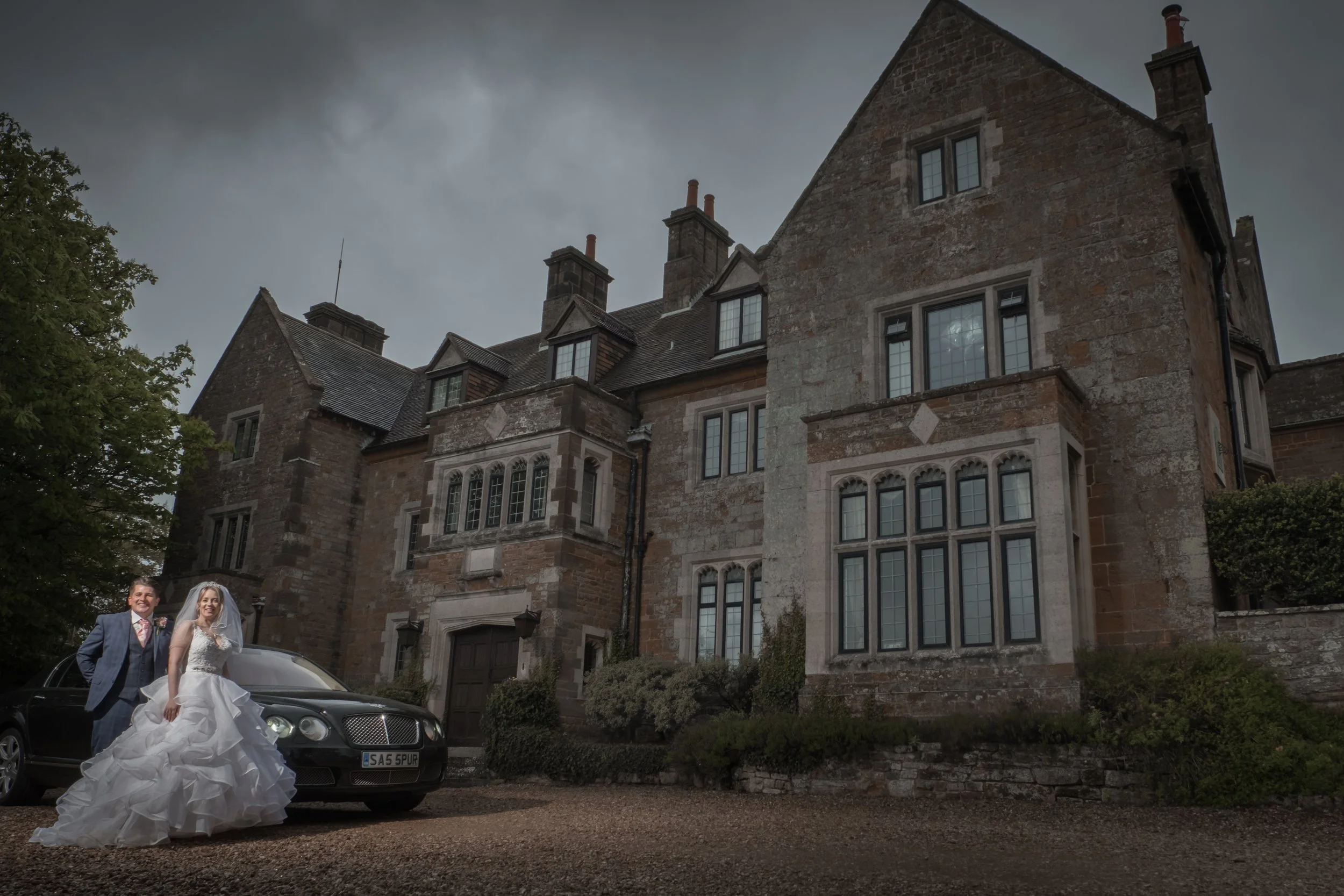 A bride and groom in wedding attire standing next to a black car in front of a large historic stone building, under a cloudy sky.