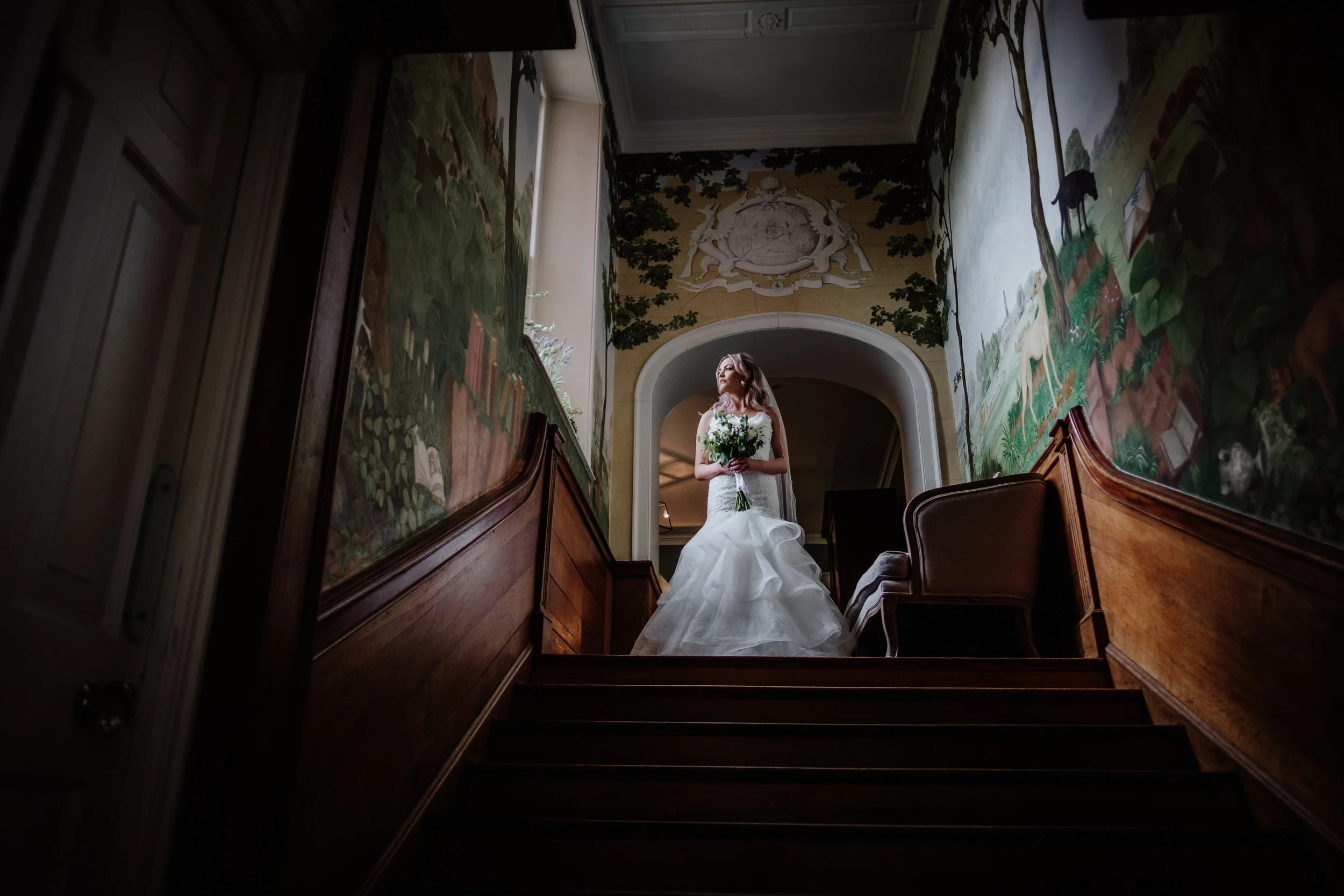 A bride in a white wedding dress holding a bouquet standing at the top of a wooden staircase in an ornately decorated hallway with painted murals and a decorative ceiling.