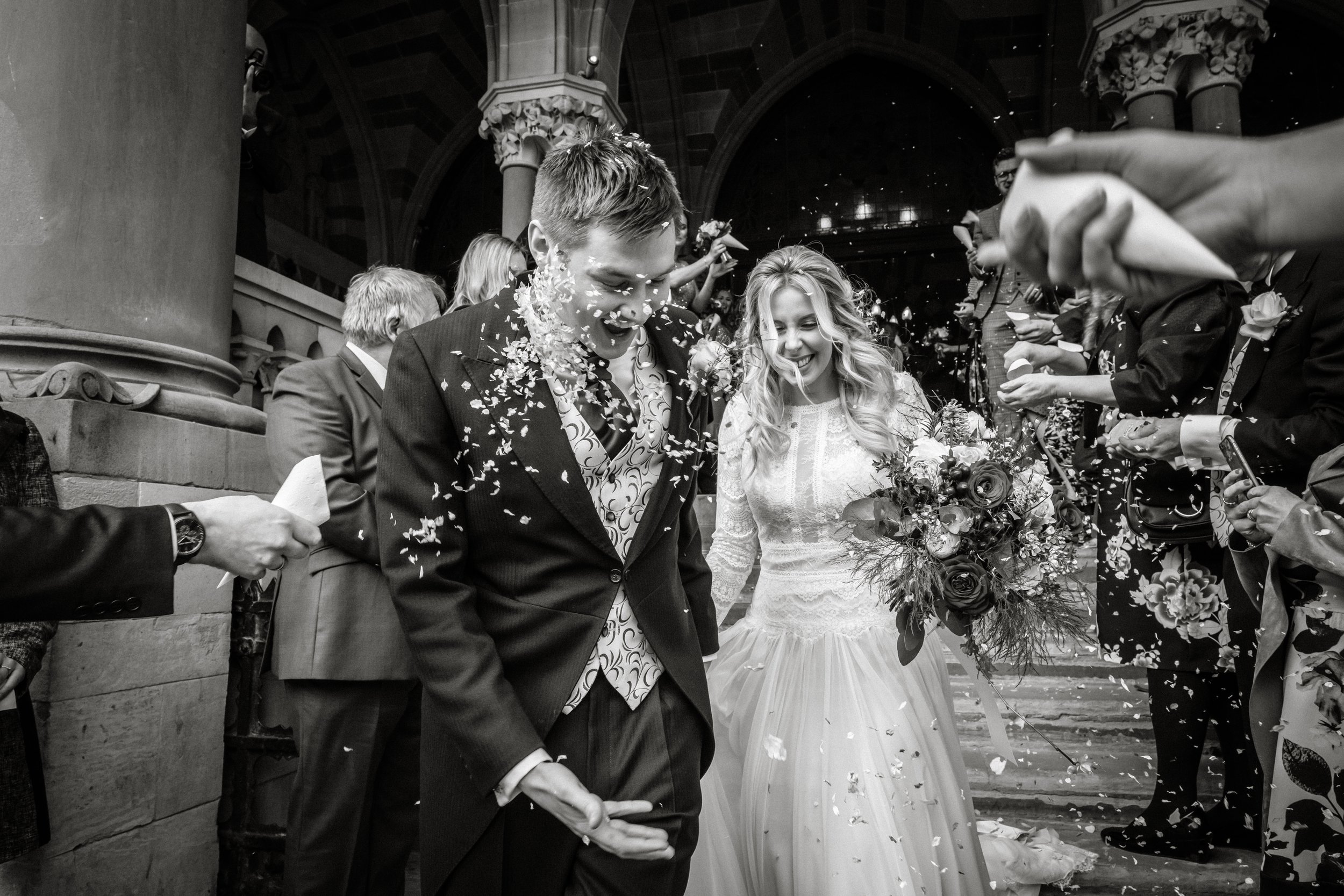 Black and white photo of a bride and groom holding hands surrounded by guests at a wedding celebration. The groom is wearing a suit with a patterned tie, and the bride is in a white lace dress holding a bouquet of flowers. Guests are throwing flower 