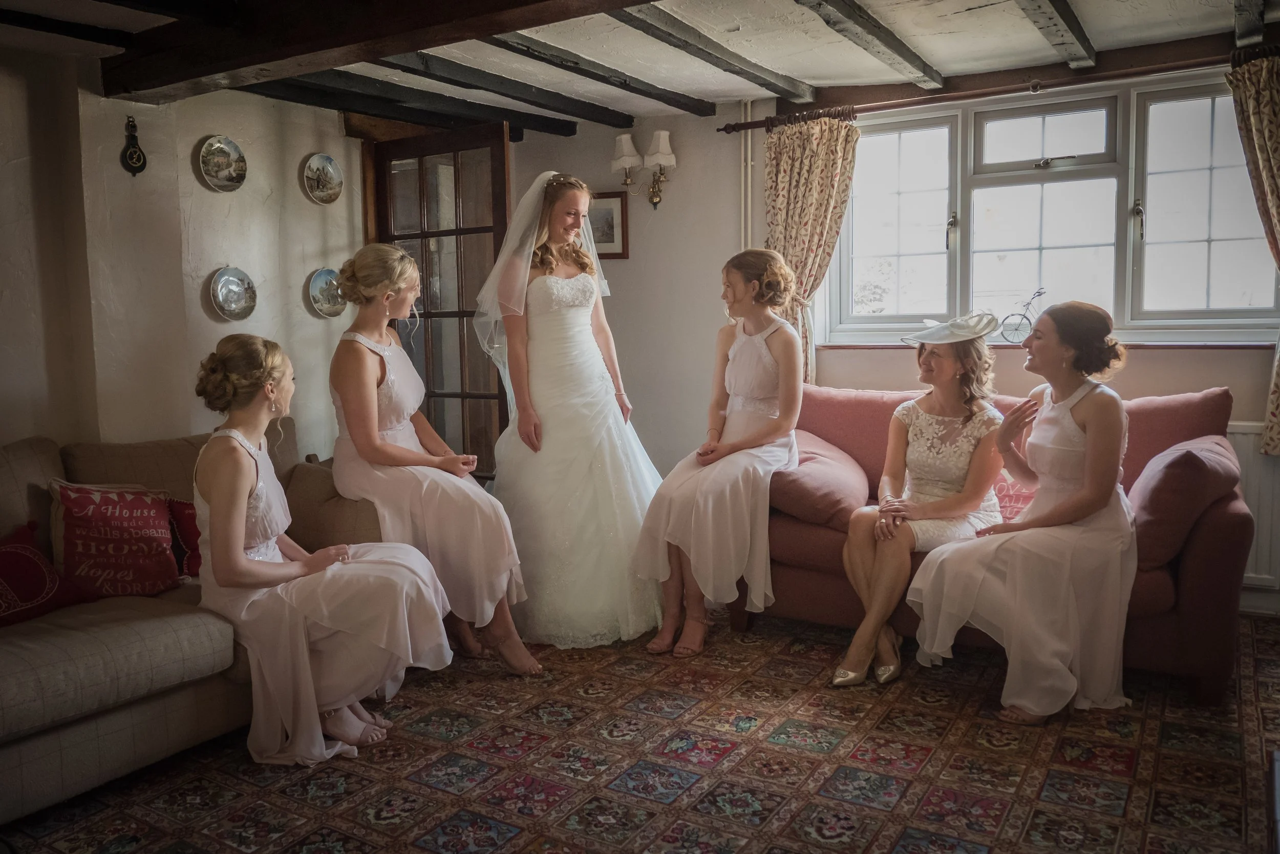 Bride in wedding dress smiling with five bridesmaids in pastel dresses sitting on a couch, in a cozy room with white walls, windows, and vintage decor.