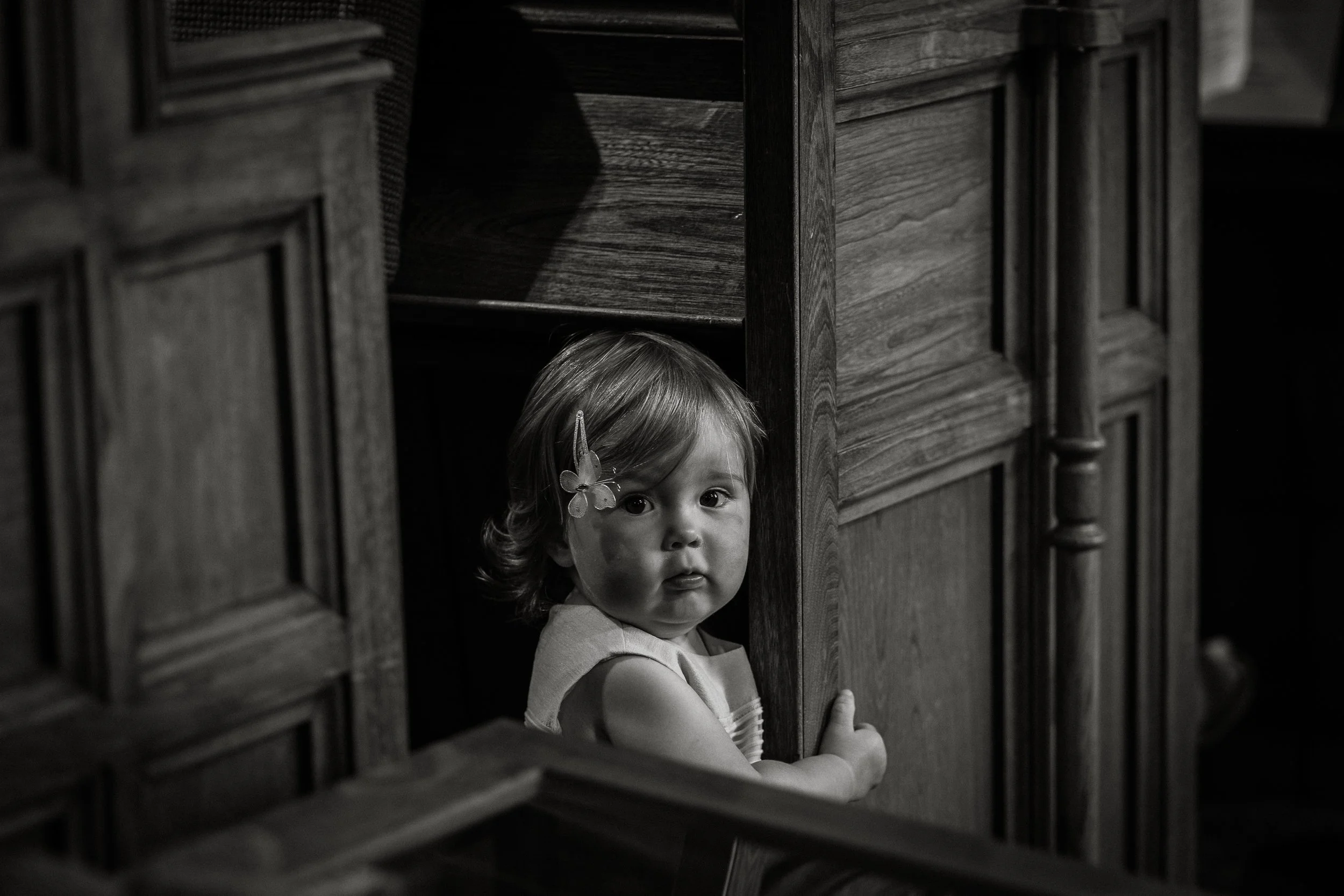 A young girl with light hair and a butterfly hair clip crouches behind wooden furniture, peeking out with wide eyes.