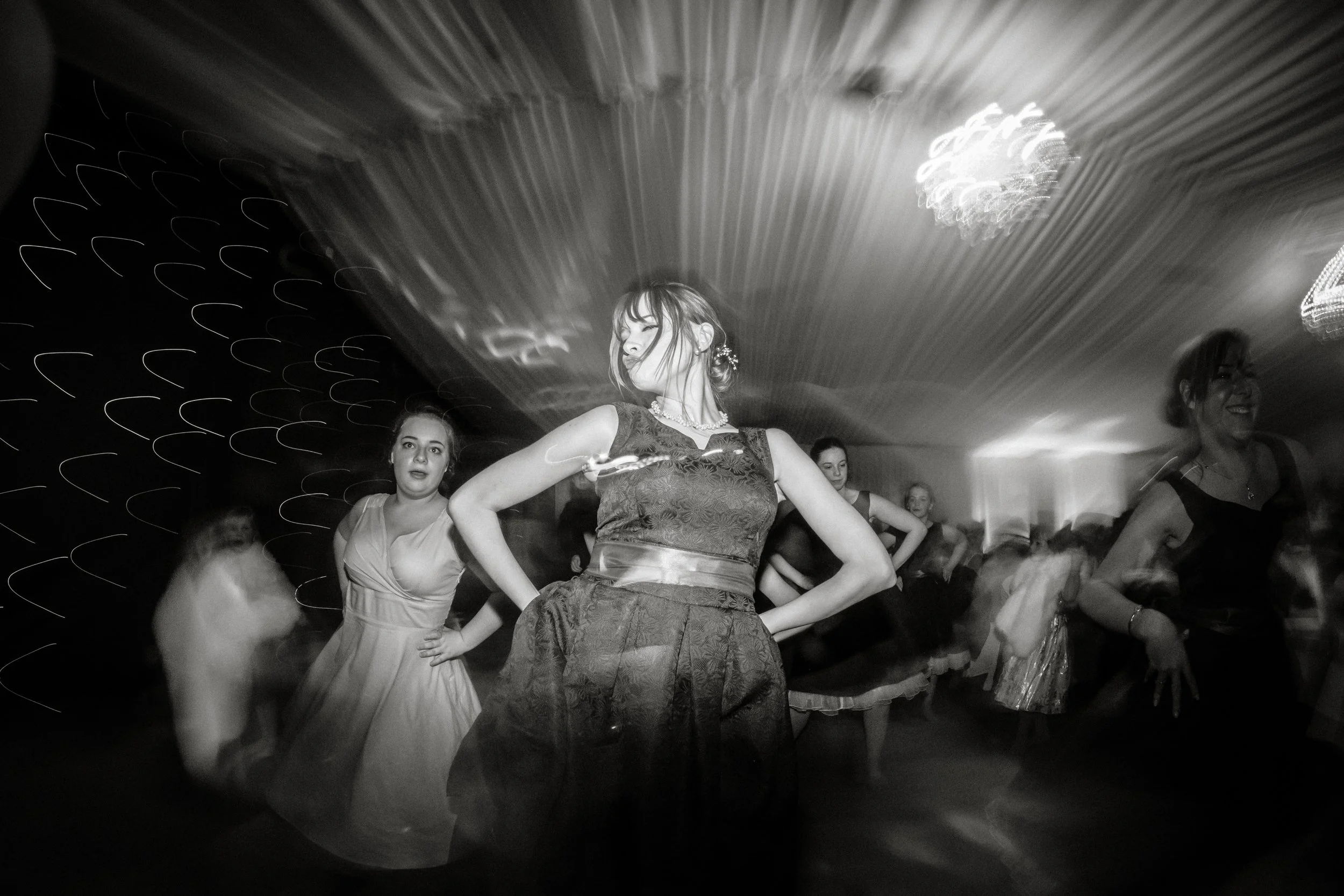 Black and white photo of women dancing at a party or celebration, with glamorous dresses and jewelry, in a room with ornate chandelier lighting.