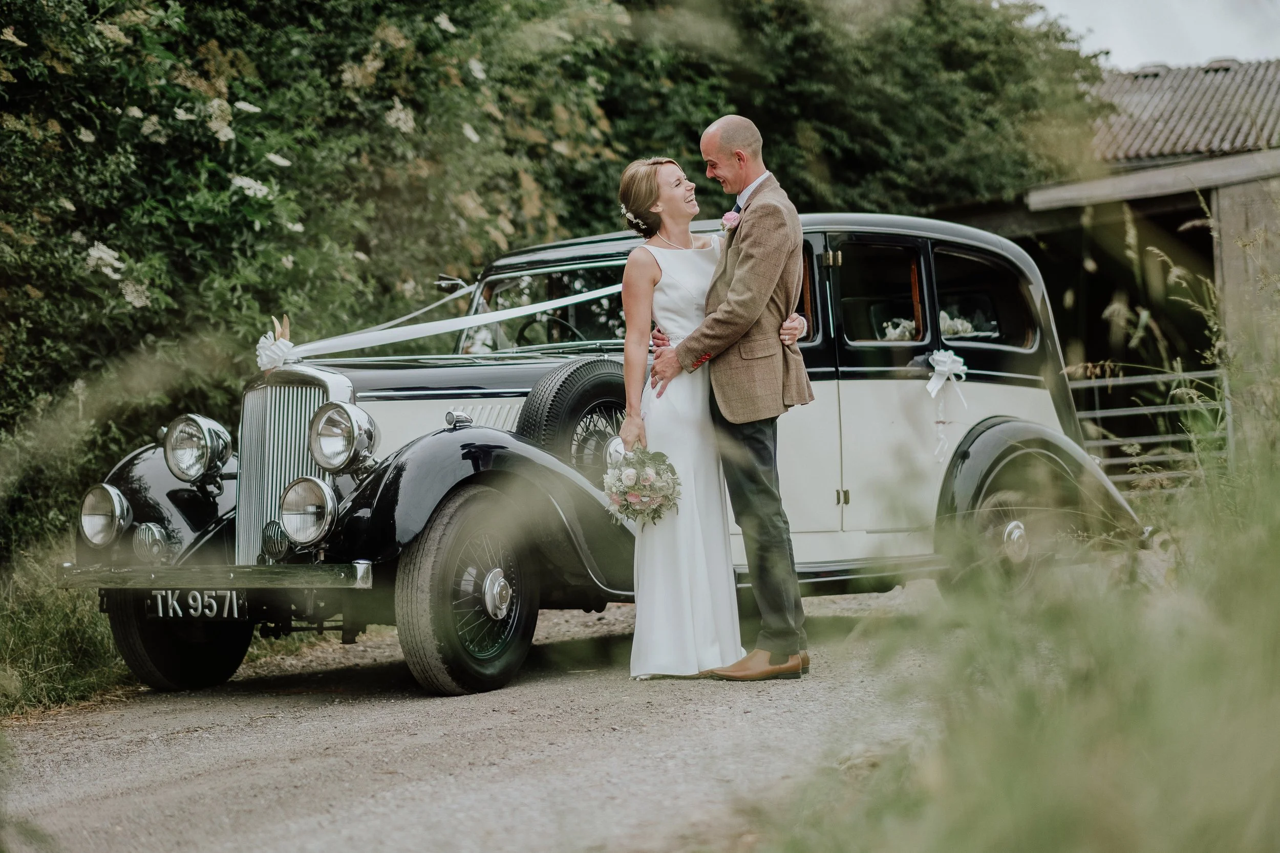 A bride and groom standing next to a vintage black and white car decorated with white ribbons and flowers, smiling and looking at each other outdoors.