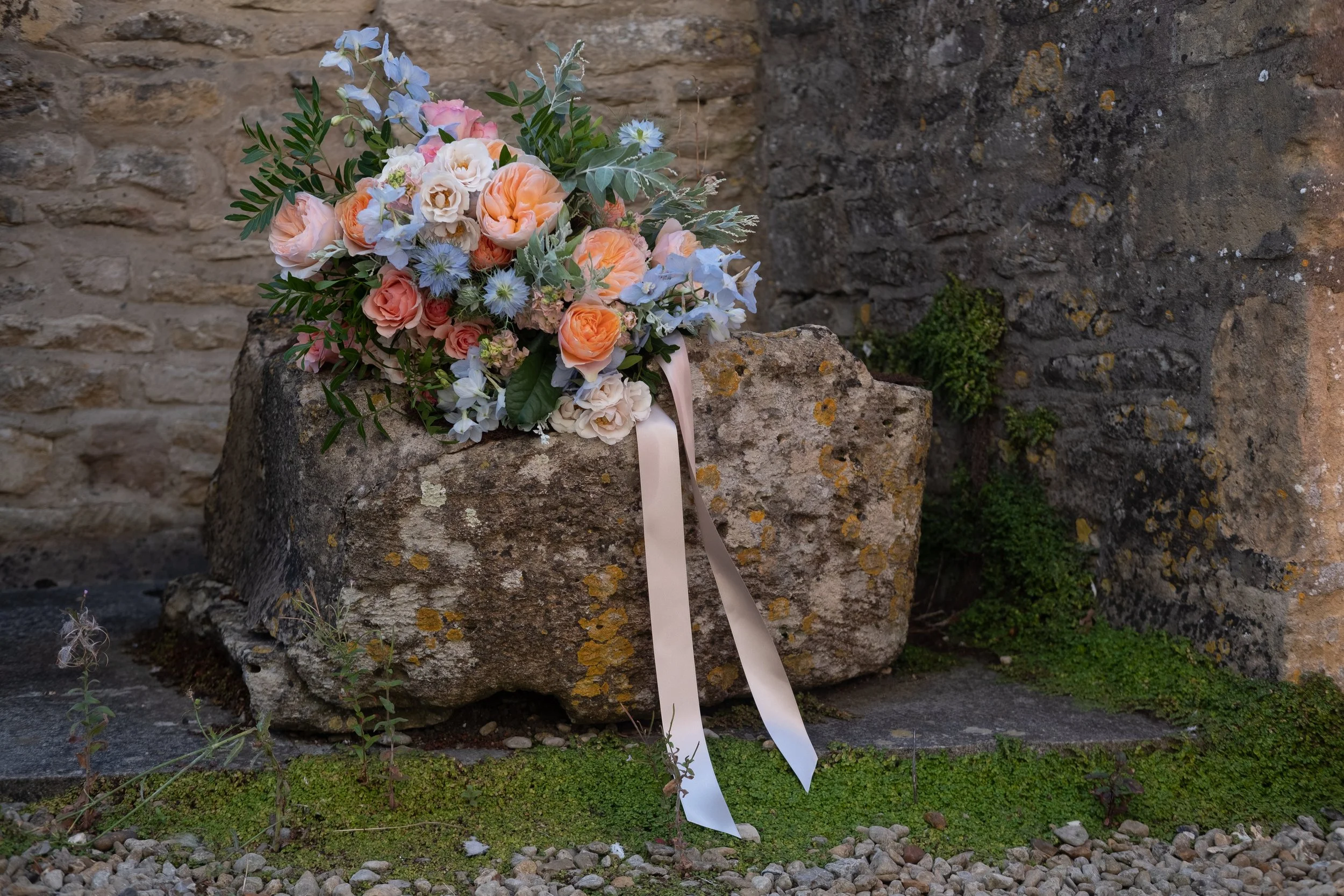 Bouquet of flowers resting on a stone block against an old brick wall, with greenery at the base.