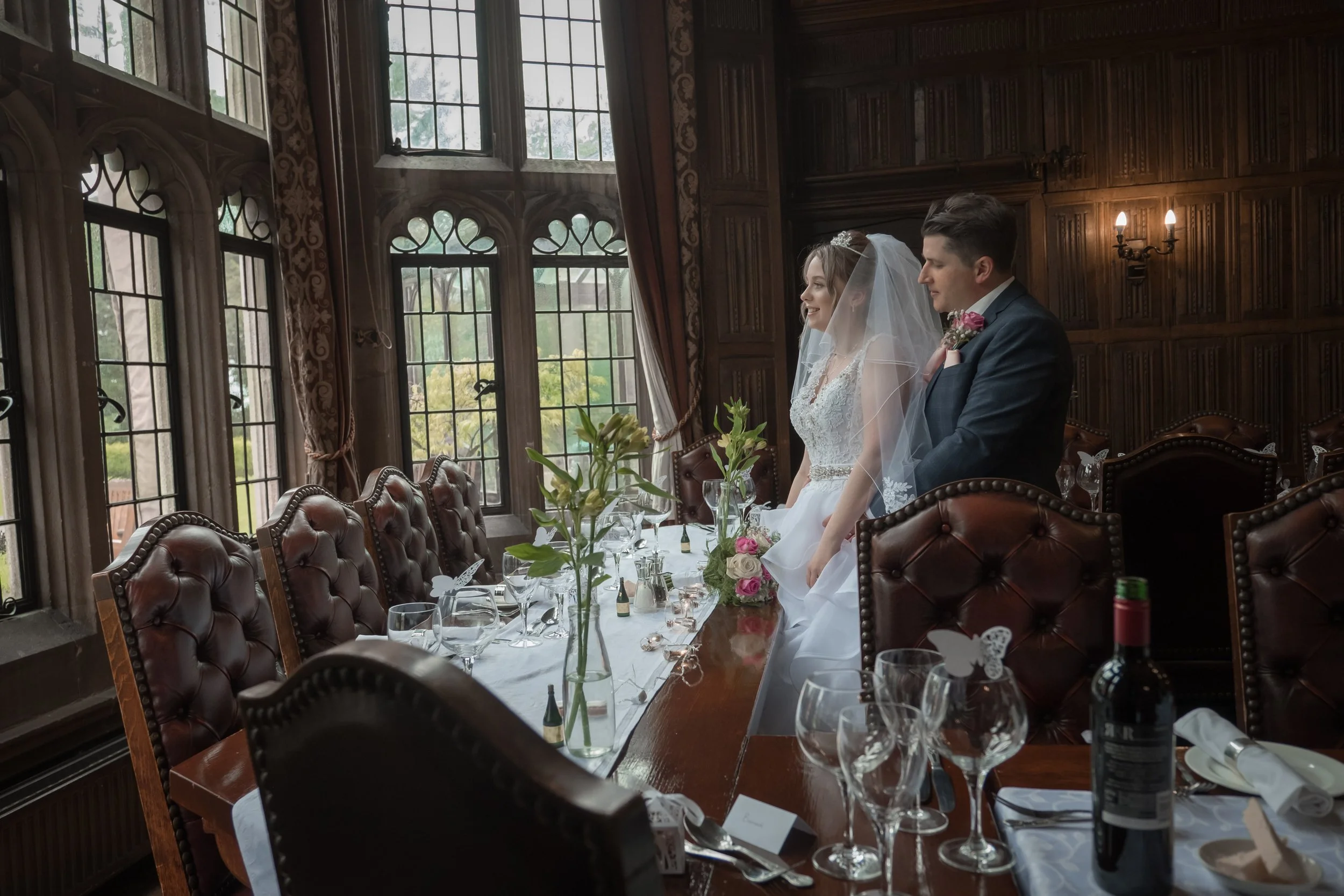 Bride and groom standing at a wedding reception table inside a wood-paneled room with large windows. The table is decorated with flowers, candles, and glasses.