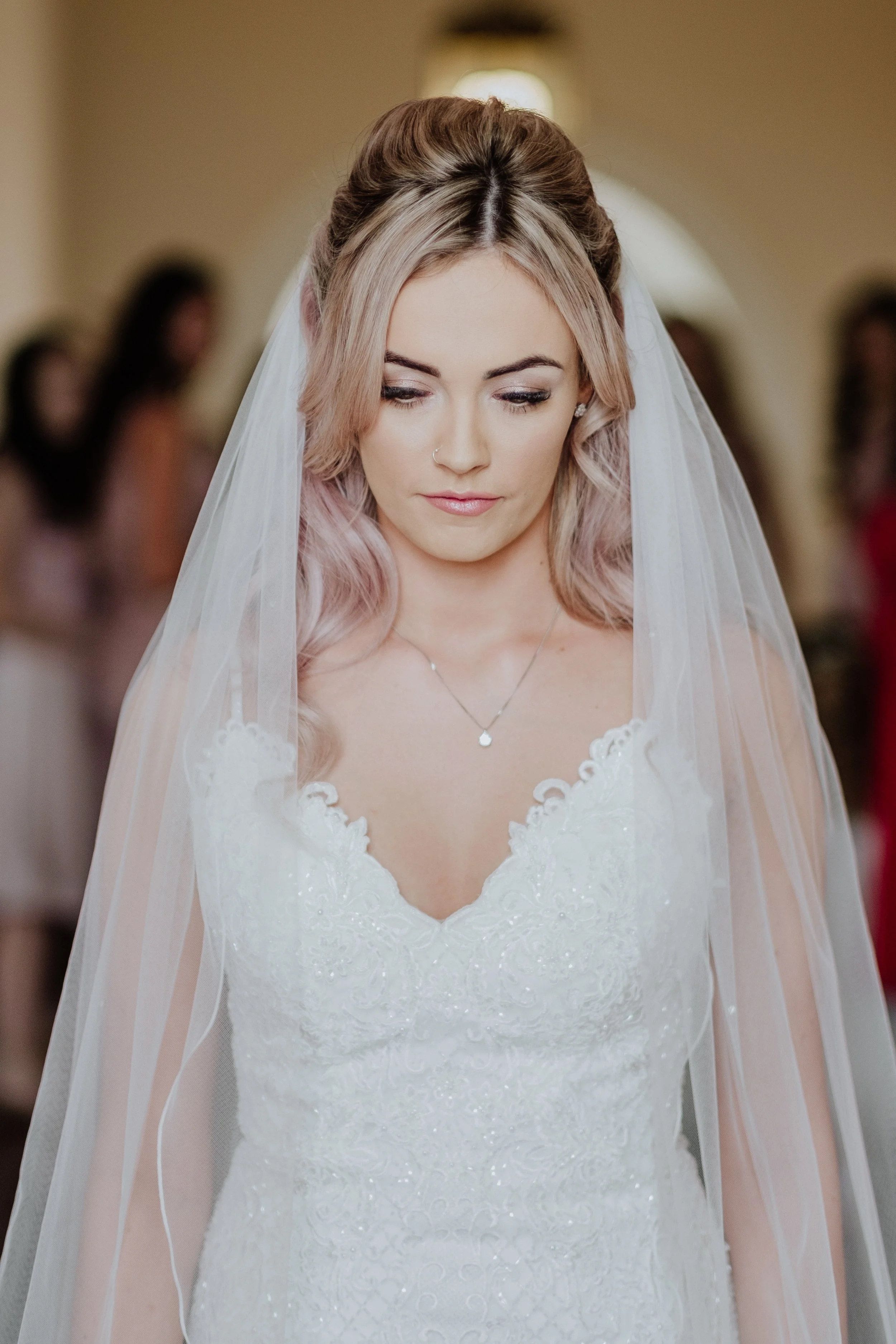 A bride with pinkish-blonde hair, wearing a white lace wedding dress and a veil, looking downward with a serene expression at a wedding ceremony.