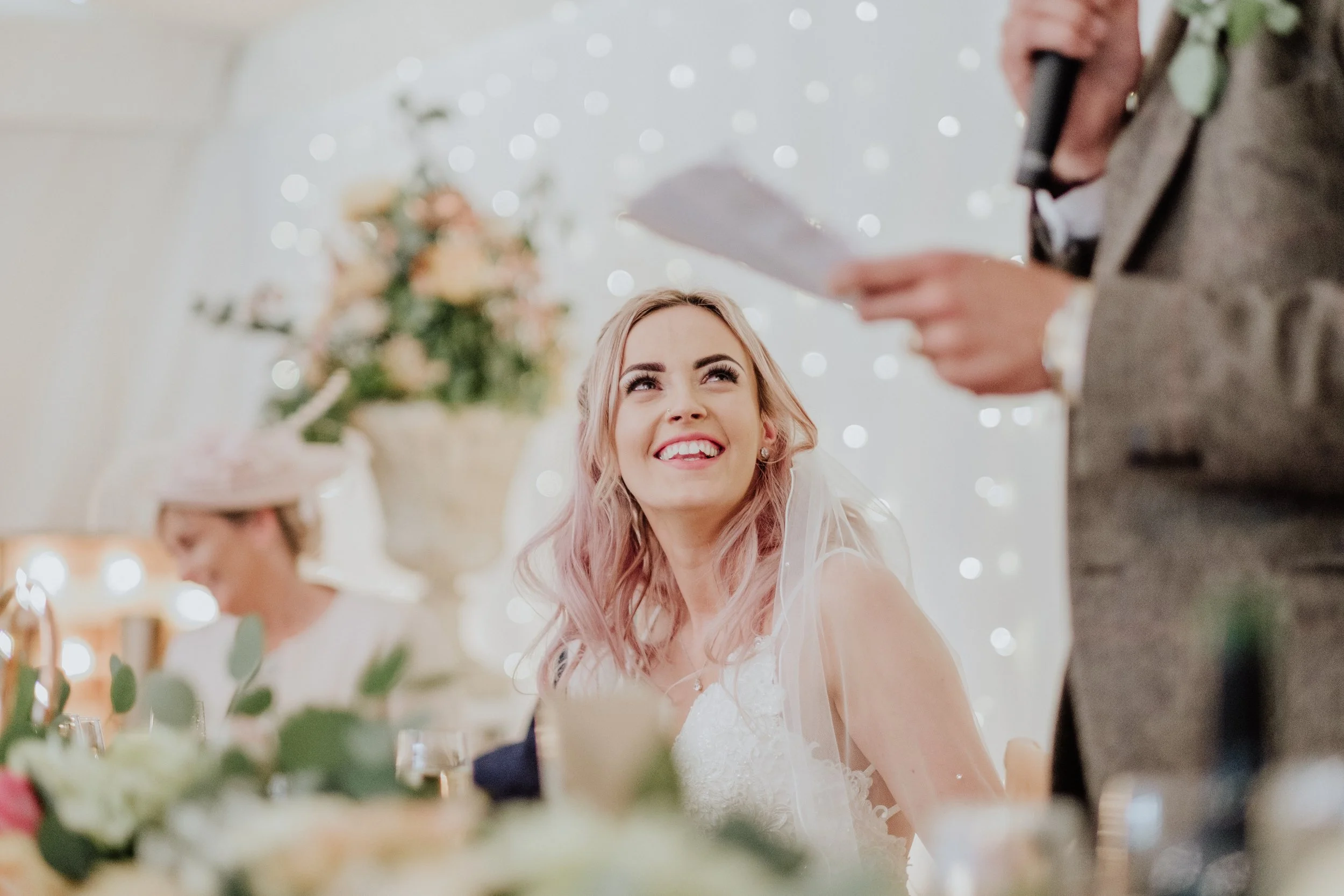 A smiling bride with pink hair looking up at her partner during a wedding reception, with floral decorations and string lights in the background.