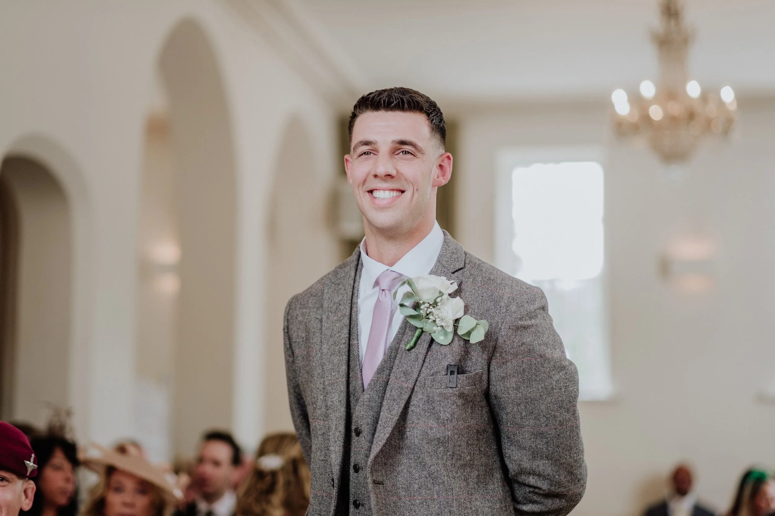 A man in a gray suit with a pink tie and a white shirt, smiling at a formal event, with a crowd of people in the background.