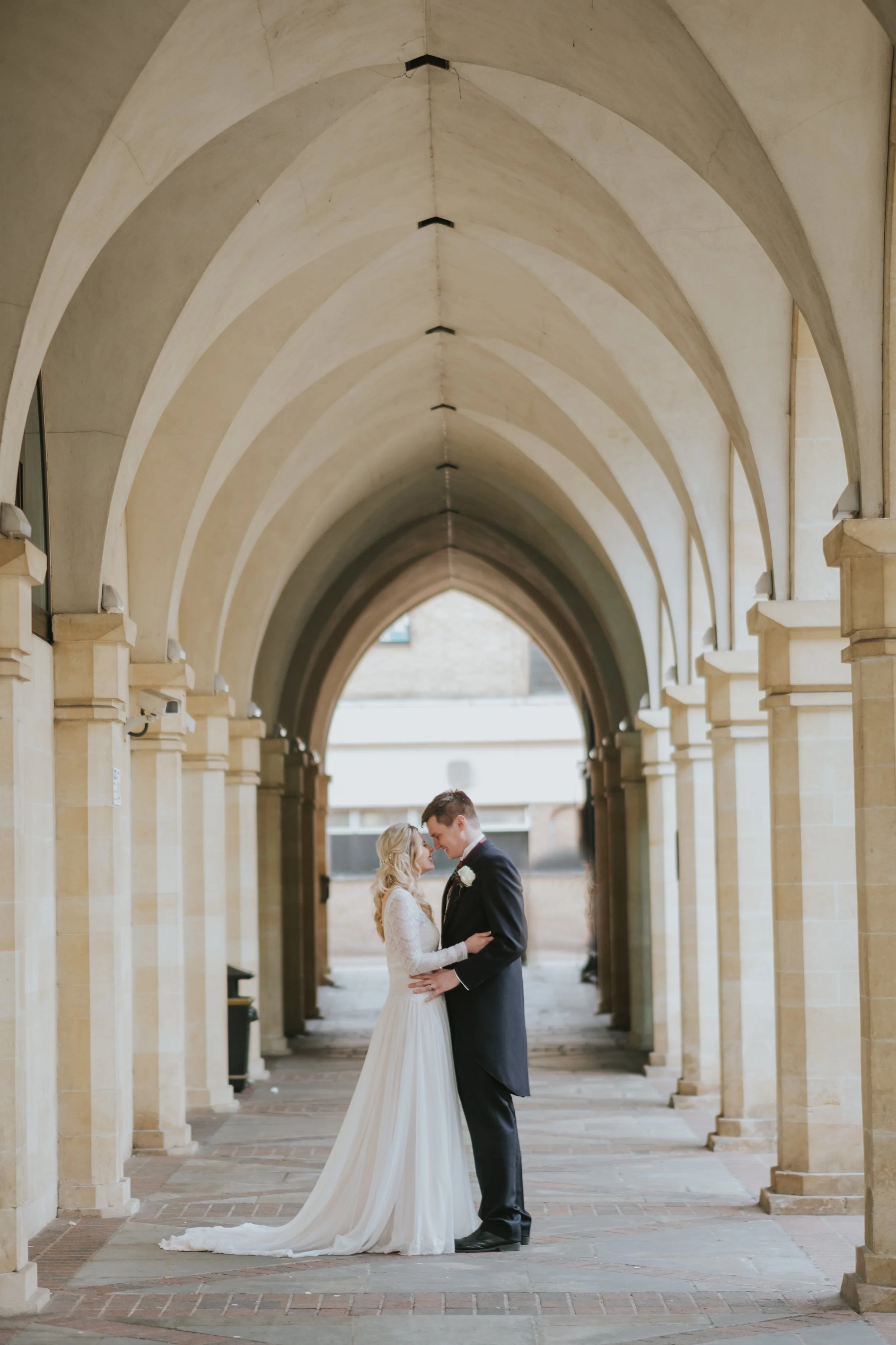 A bride and groom standing close together under a stone arched walkway, gazing into each other's eyes, in a romantic wedding setting.