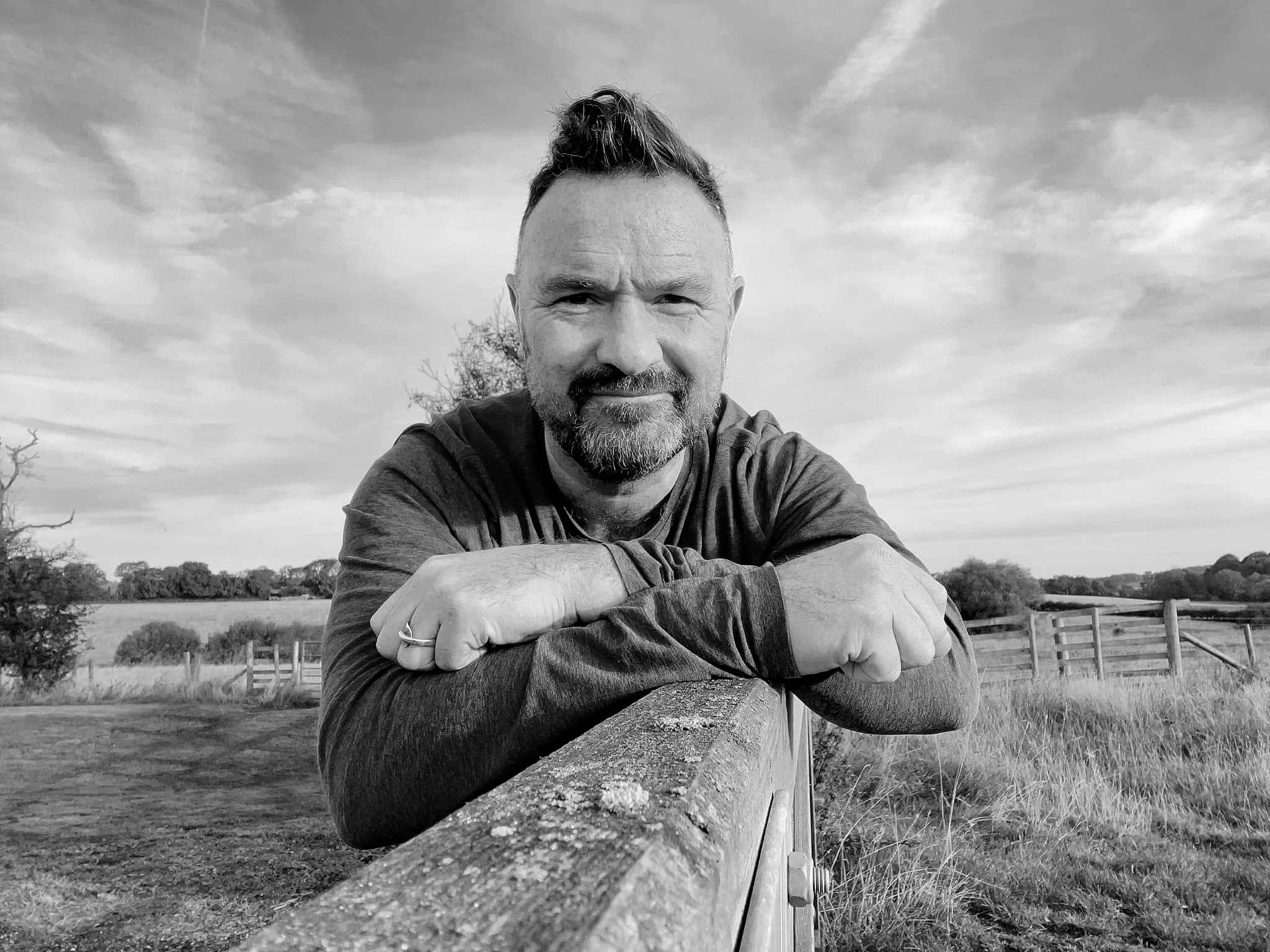 A man with a beard and styled hair leaning on a wooden railing outdoors, with trees, a fence, and a cloudy sky in the background, in black and white.