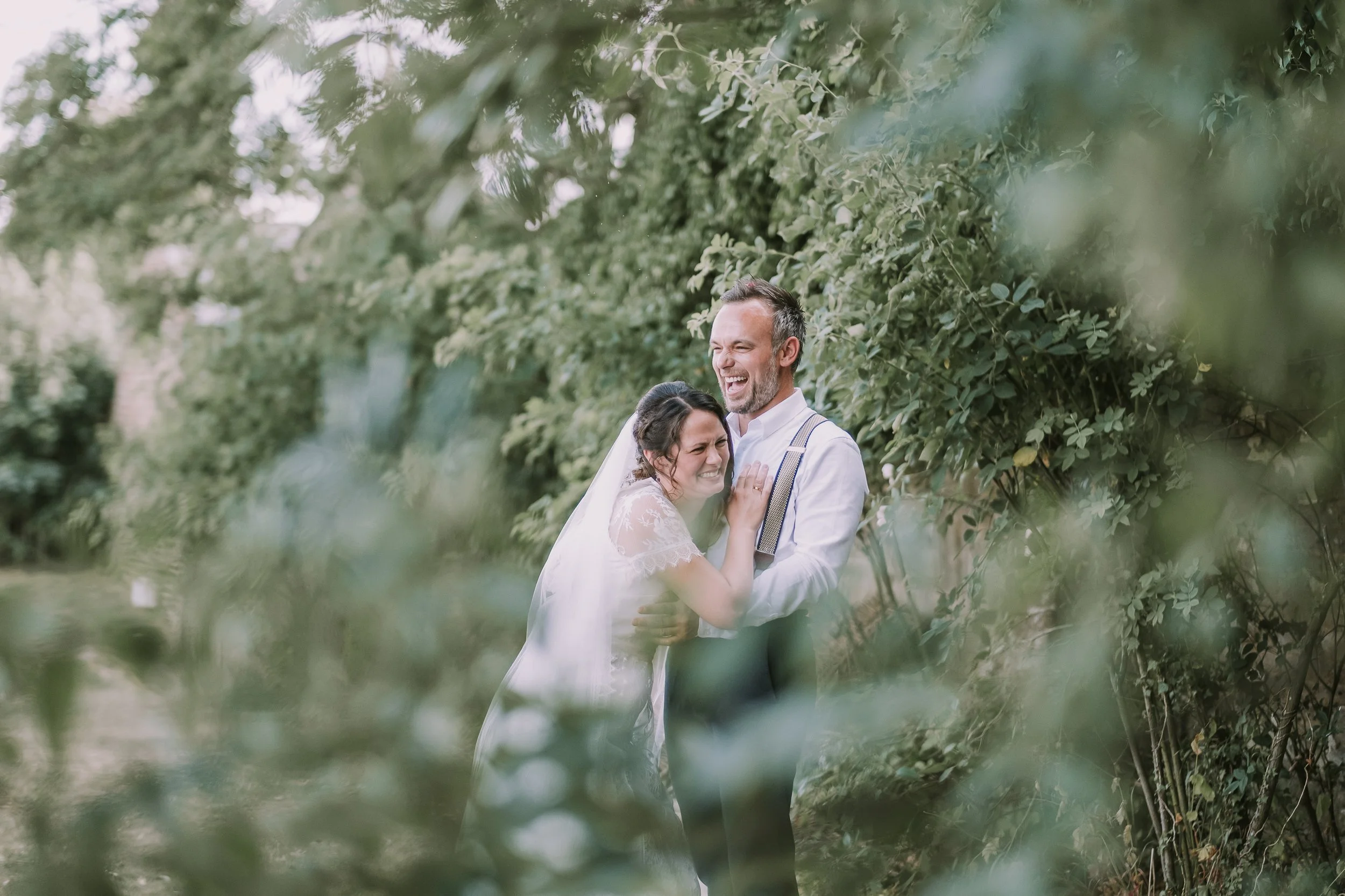 A happy couple, a man and a woman, embracing each other outdoors amidst green foliage, smiling and laughing.