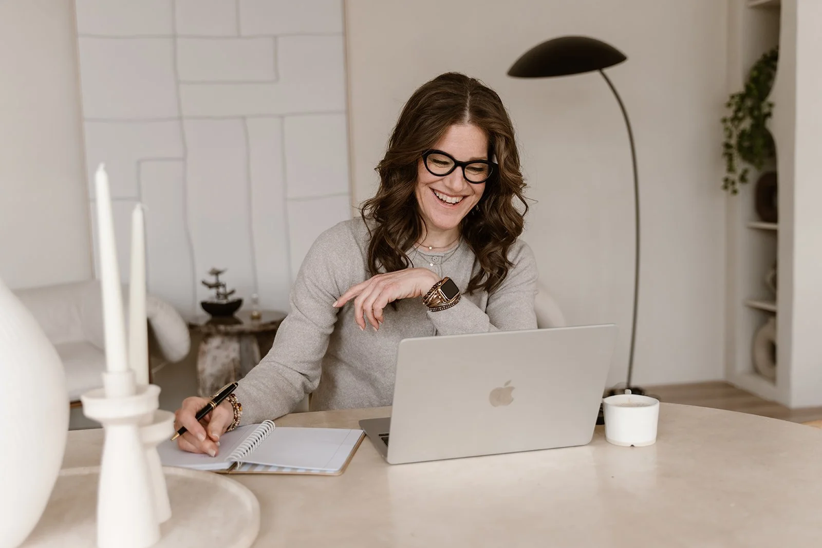 A woman with brown wavy hair, glasses, and a gray sweater, sitting at a desk working on a silver Apple laptop while smiling. She holds a pen and has a notebook open in front of her. There are white candles and a cup on the desk, and a lamp, potted plant, and shelves in the background.