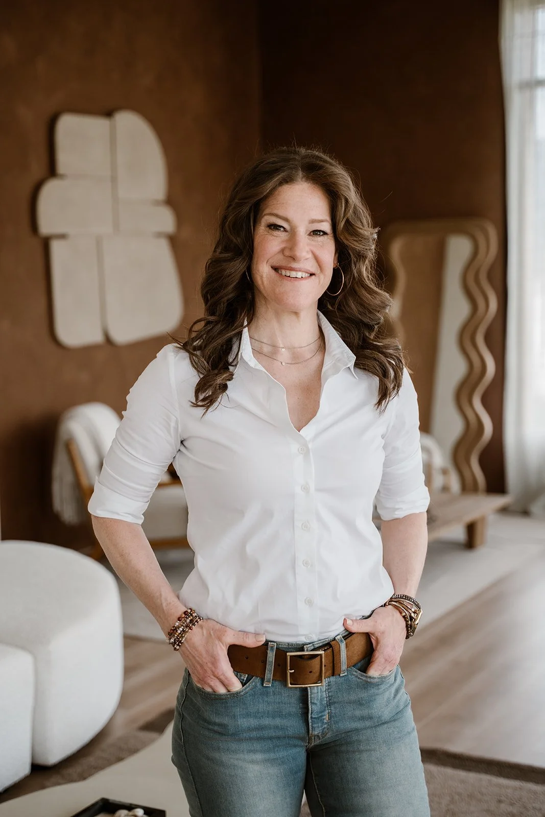 A woman with brown wavy hair smiling, wearing a white button-up shirt and blue jeans, standing in a warmly decorated living room with modern art and a large window.