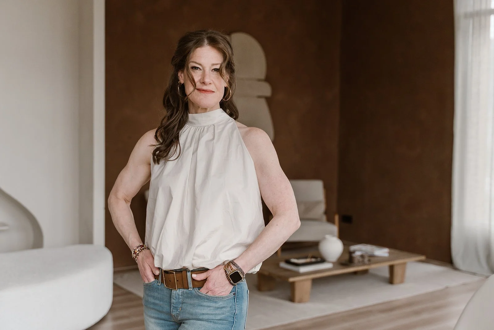 A woman with brown hair wearing a white sleeveless blouse and blue jeans standing in a modern living room.