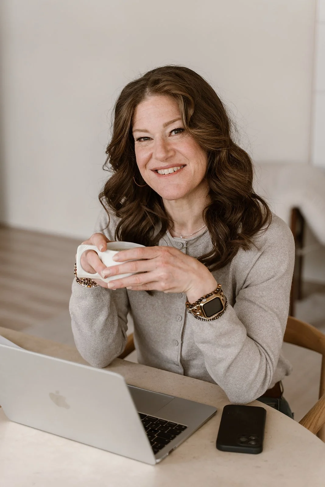 A woman with wavy brown hair smiling, holding a white mug, sitting at a table with a silver MacBook laptop and a black smartphone.