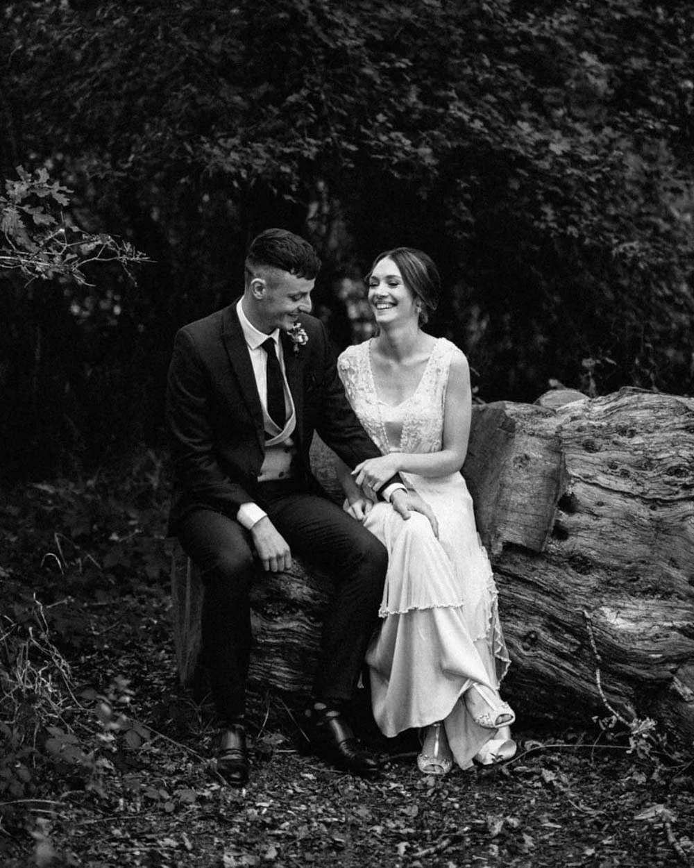 black and white photo of bride and groom sitting on tree stump smiling and looking towards each other