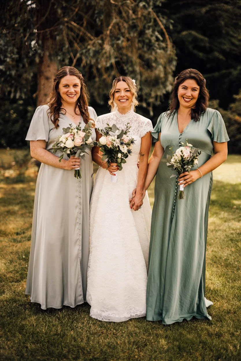 bride with two bridesmaids wearing muted green dresses smiling to camera holding bouquets