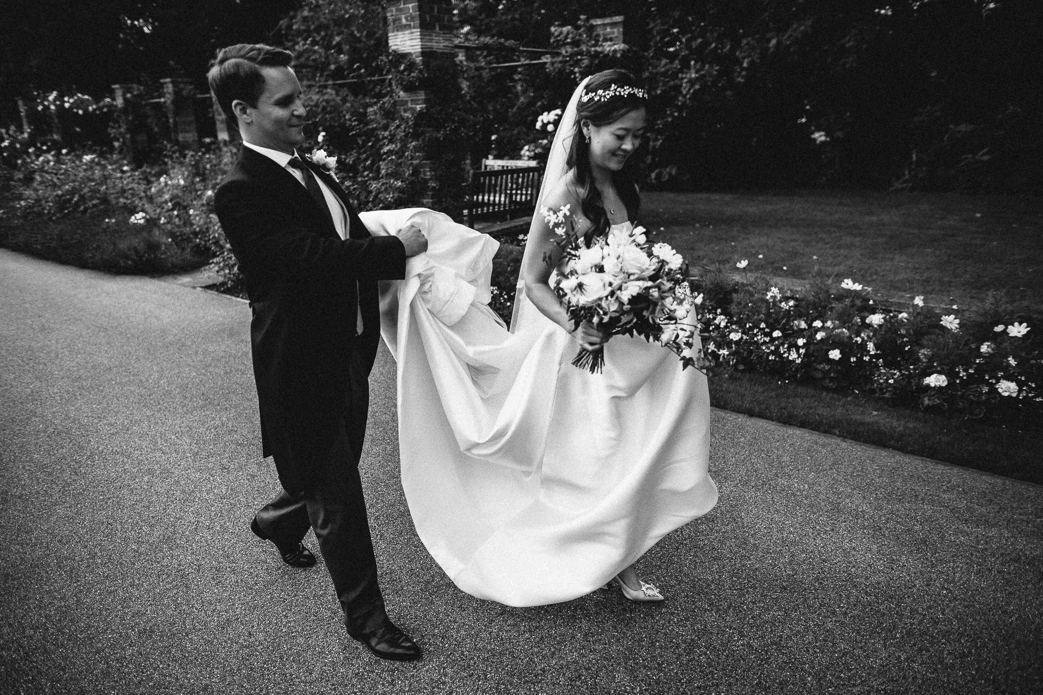  Bride and Groom walk through the grounds of the Hurlingham Club as the Groom lifts the bride’s dress 