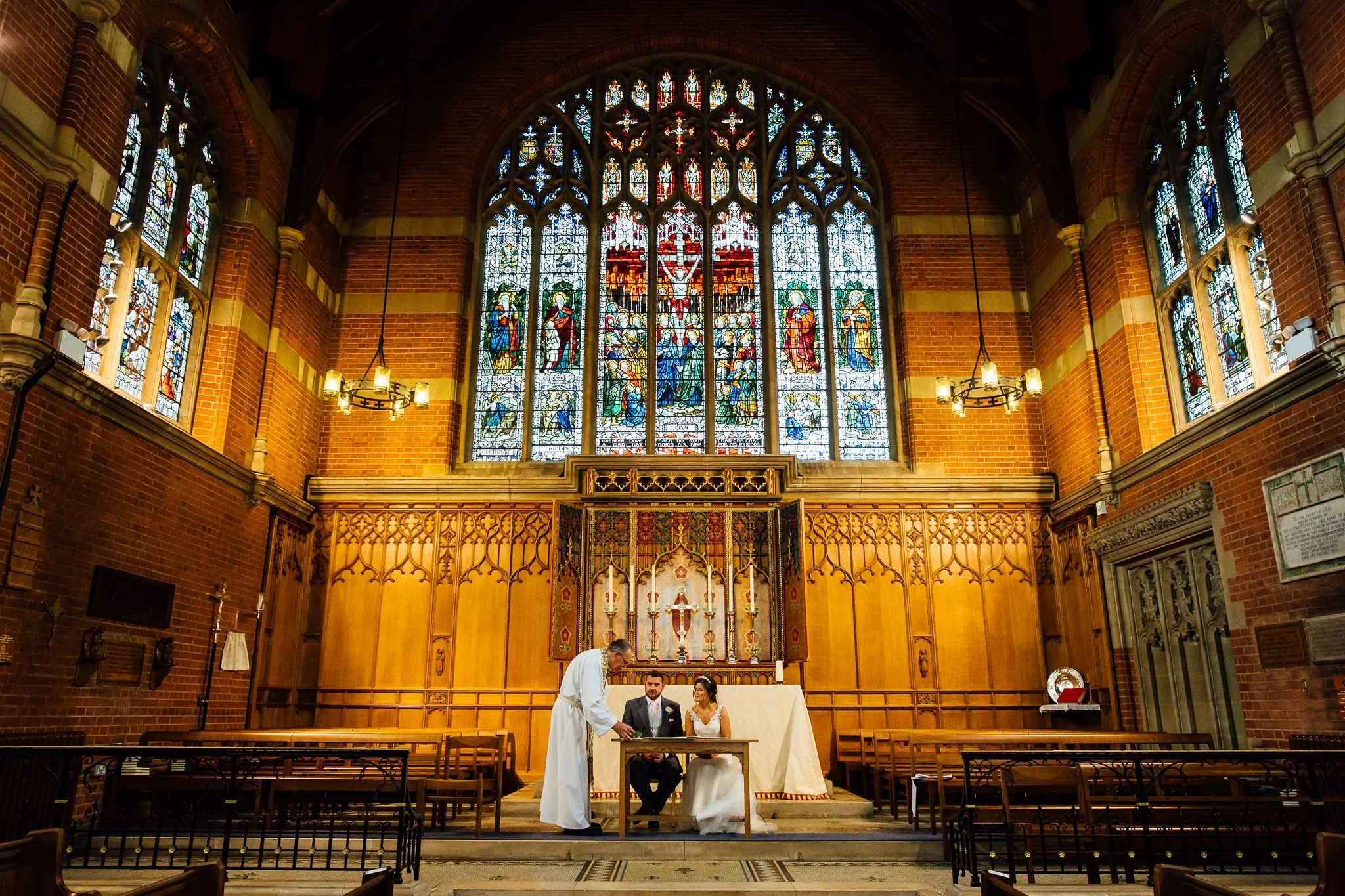  Bride and Groom are seated to sign the marriage certificate in front of a large stain glassed window  at the Chapel of St Luke at Epsom College 