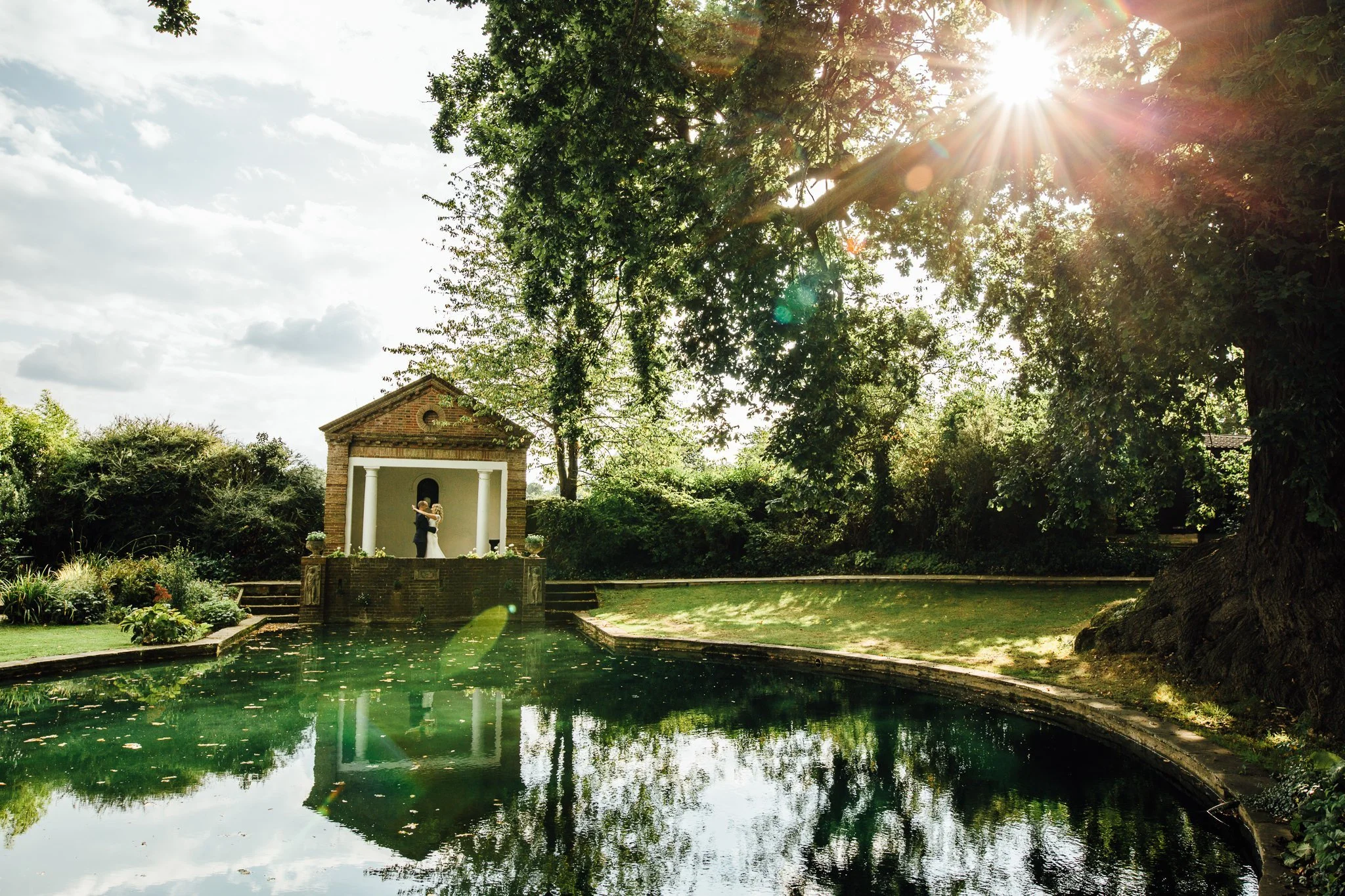  Bride and Groom stand in front of the temple at Micklefield Hall as the sun shines through the tress 