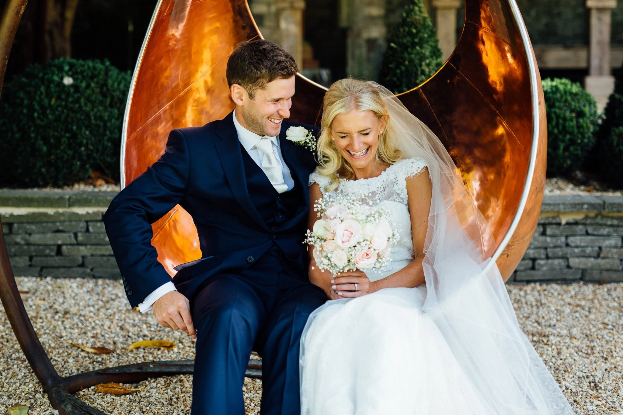  Bride and groom sit on a chair suspended off the ground at Aynhoe Park 