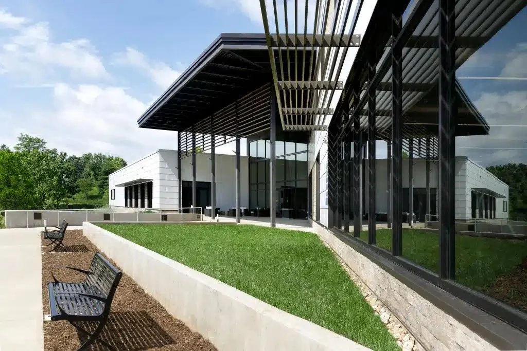 Modern building with large glass windows, metal awnings, and a landscaped area with benches, surrounded by greenery.