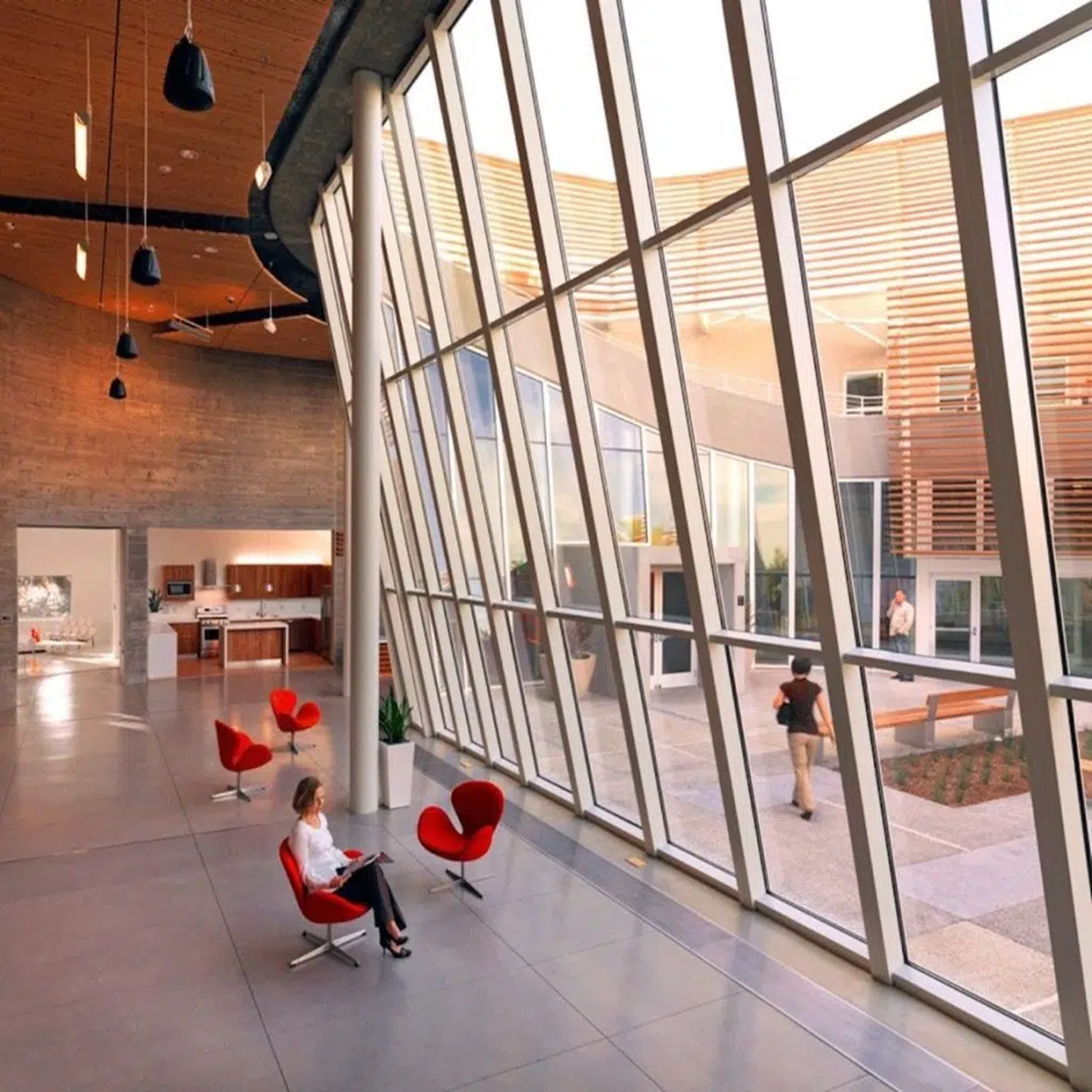 Interior of a modern building with large glass windows, wooden ceiling, and brick walls, featuring red chairs and a few people inside.