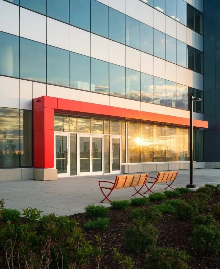 Modern office building with reflective glass windows and a red entrance canopy, two benches, a lamppost, and landscaped bushes at sunset.