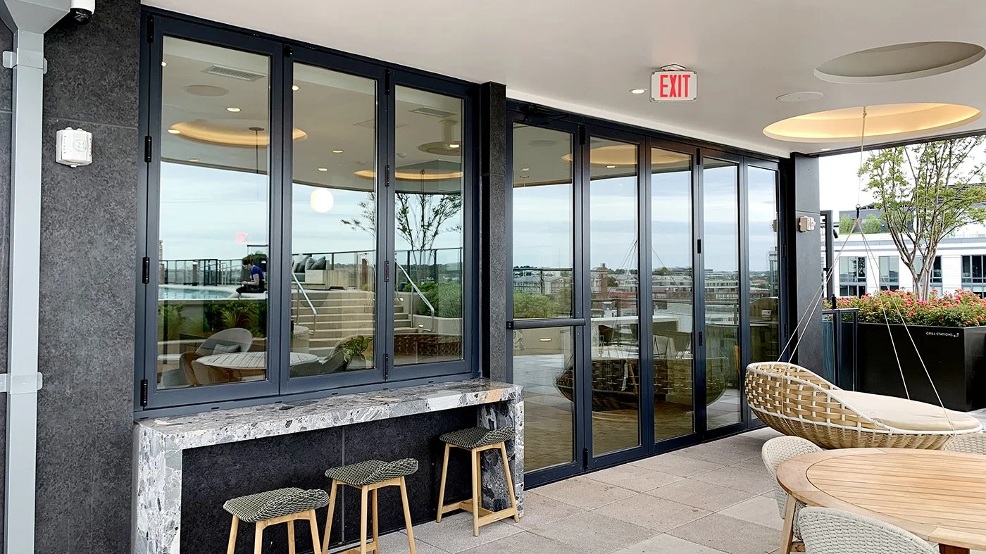 Outdoor seating area with wicker chairs and a round wooden table, glass sliding doors leading to an indoor space, and potted plants, in an urban rooftop setting.