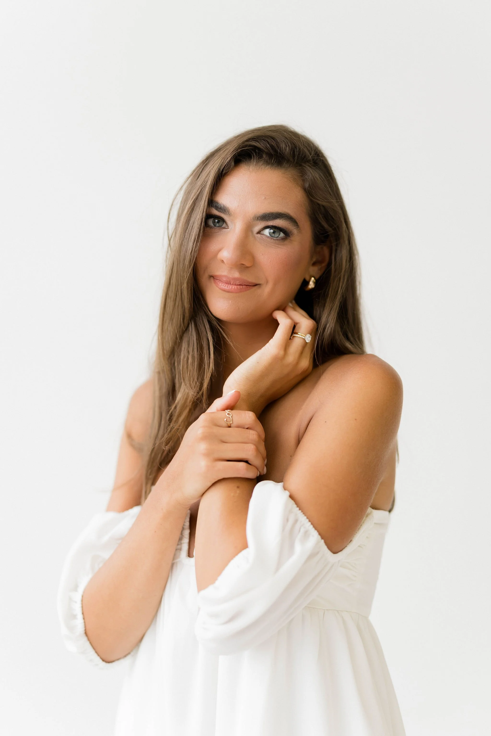 A young woman with long brown hair and blue eyes, wearing a white off-shoulder dress and gold jewelry, smiling softly against a plain white background.