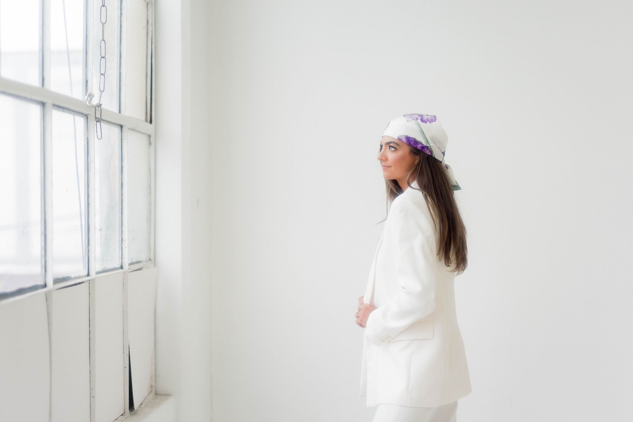 A woman wearing a white headscarf with purple floral patterns and a white coat, standing indoors near a window with metal bars, looking out thoughtfully.